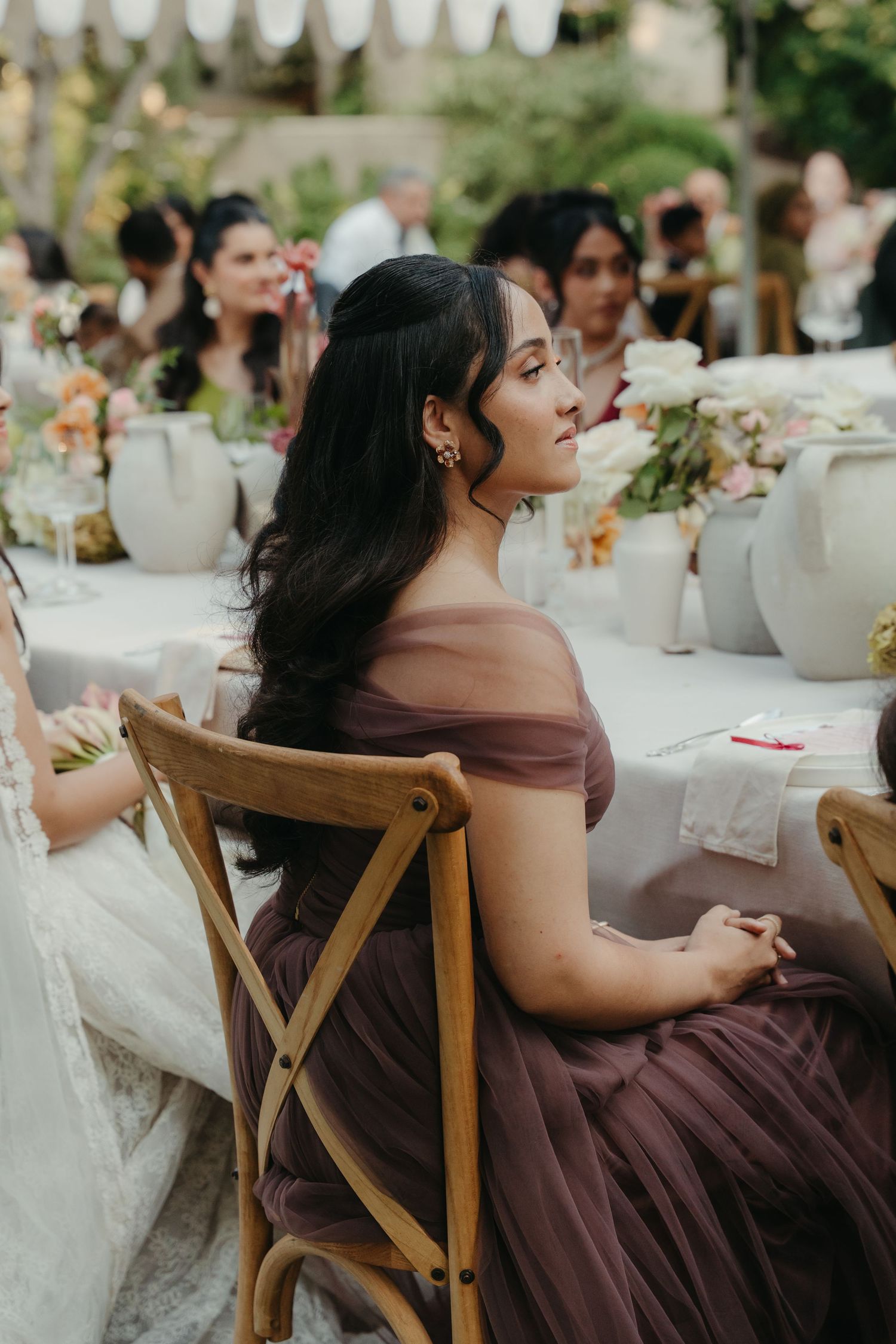 A bridesmaid in a mauve off-shoulder tulle gown sits gracefully at an outdoor wedding reception table.