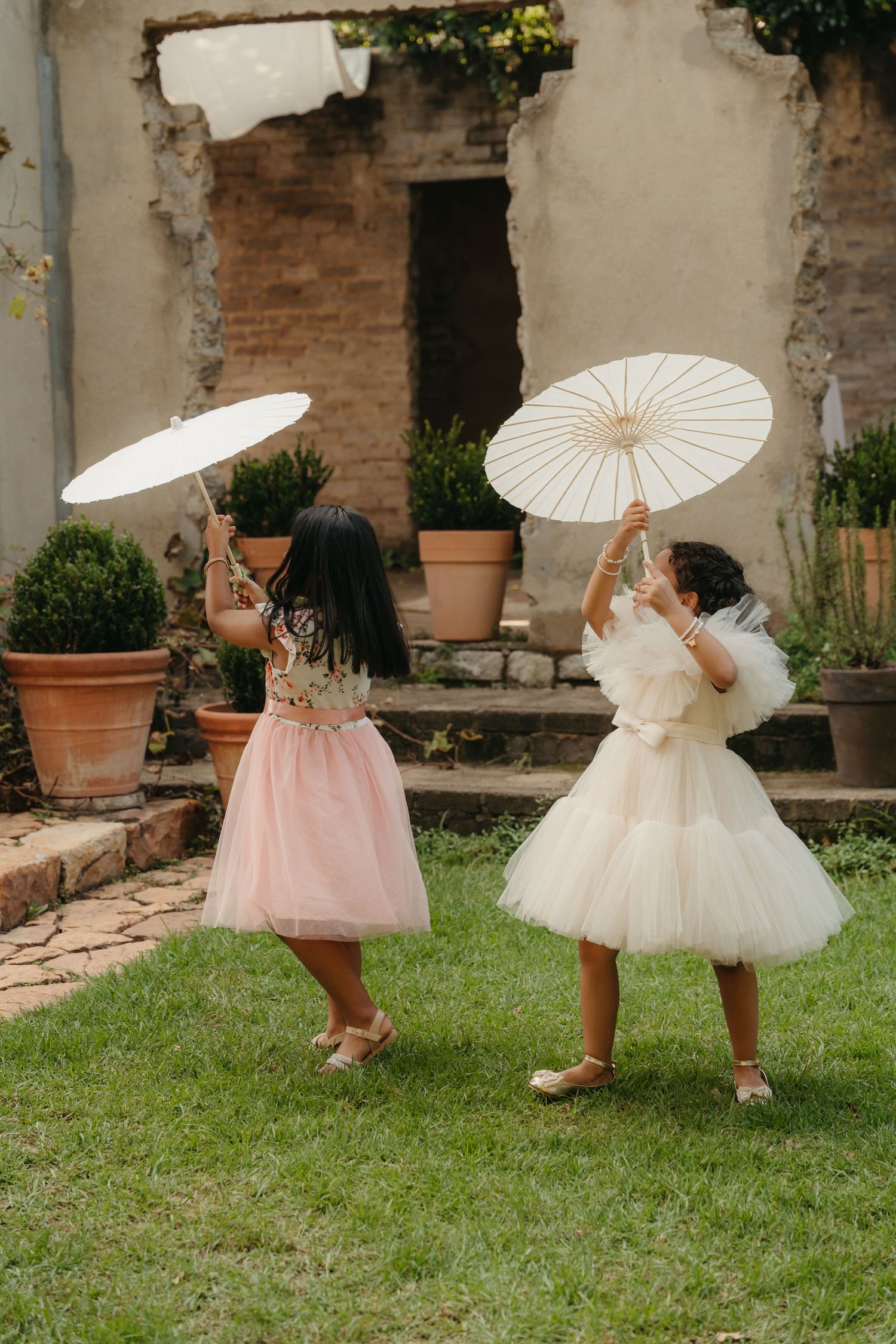 Two flower girls in tulle dresses twirl white parasols on green grass at an outdoor garden wedding venue.