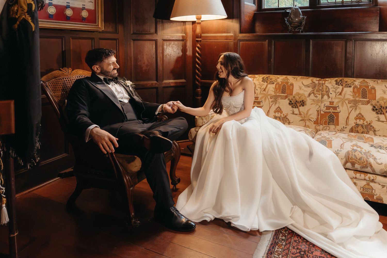 Bride and groom holding hands in an elegant wood-paneled room, seated on vintage furniture in wedding attire.