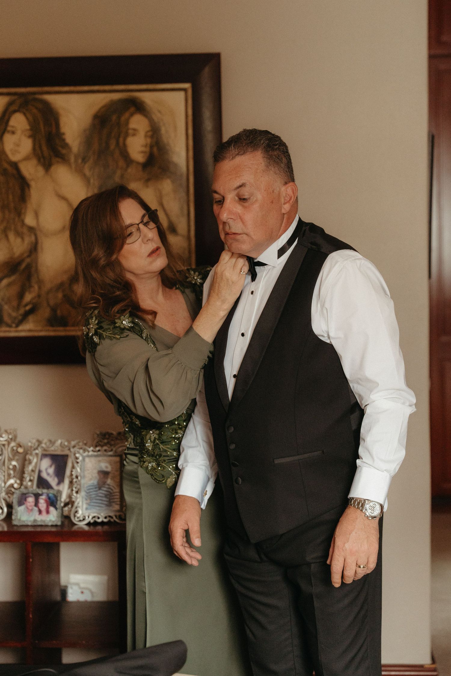 A woman in a green dress adjusts a man's bow tie as he prepares in formal attire for a wedding.