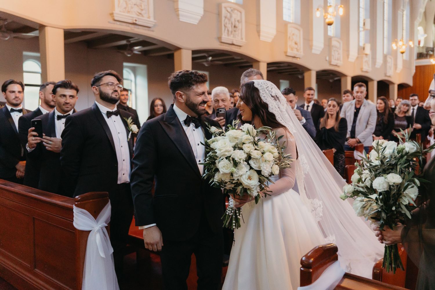 Bride and groom walk down church aisle smiling, bride holding white floral bouquet, groomsmen in black tuxedos standing nearby.