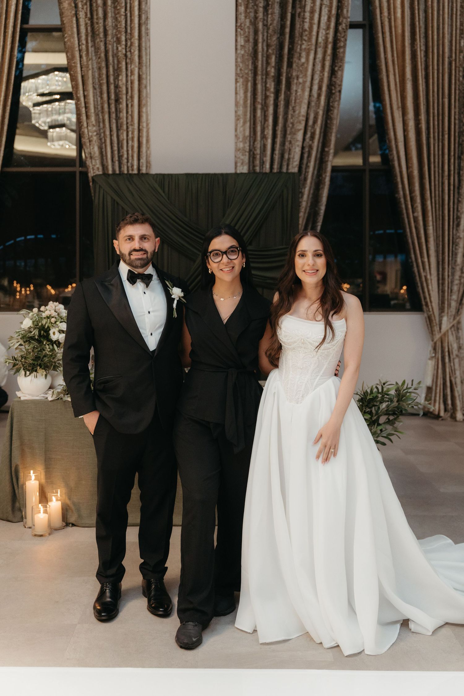 Bride and groom in wedding attire pose with officiant in black jumpsuit before elegant candlelit ceremony backdrop.