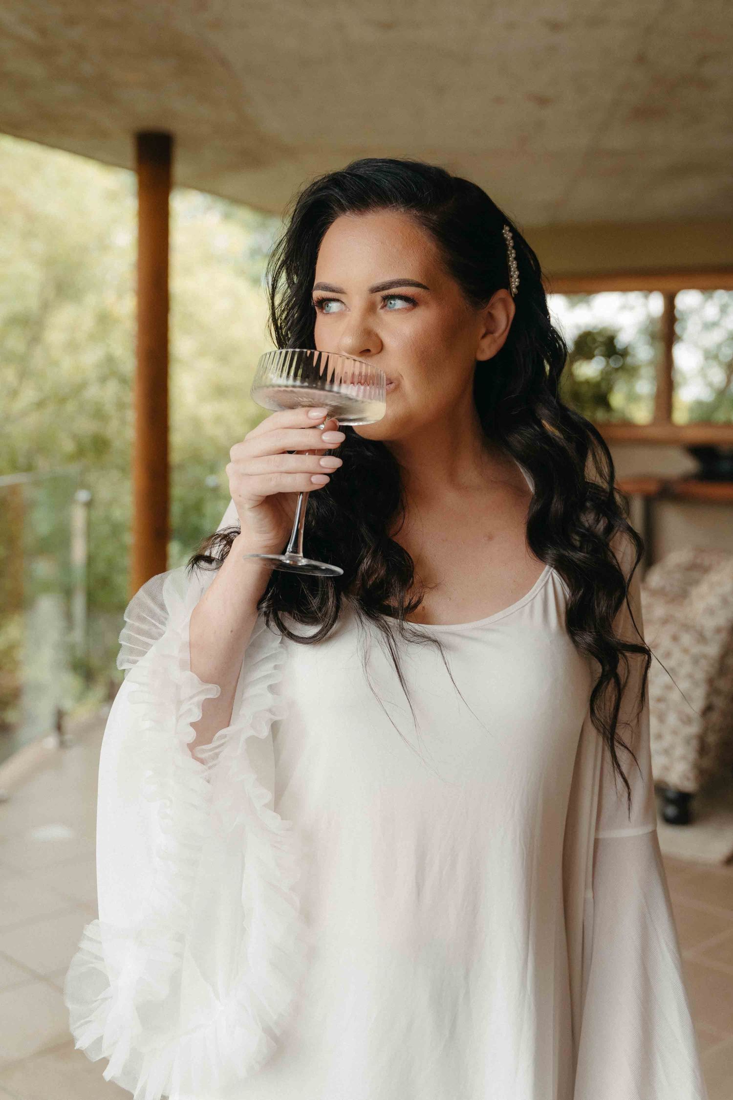 A bride in a white ruffled gown sips champagne from a coupe glass, her dark wavy hair adorned with a pearl clip.
