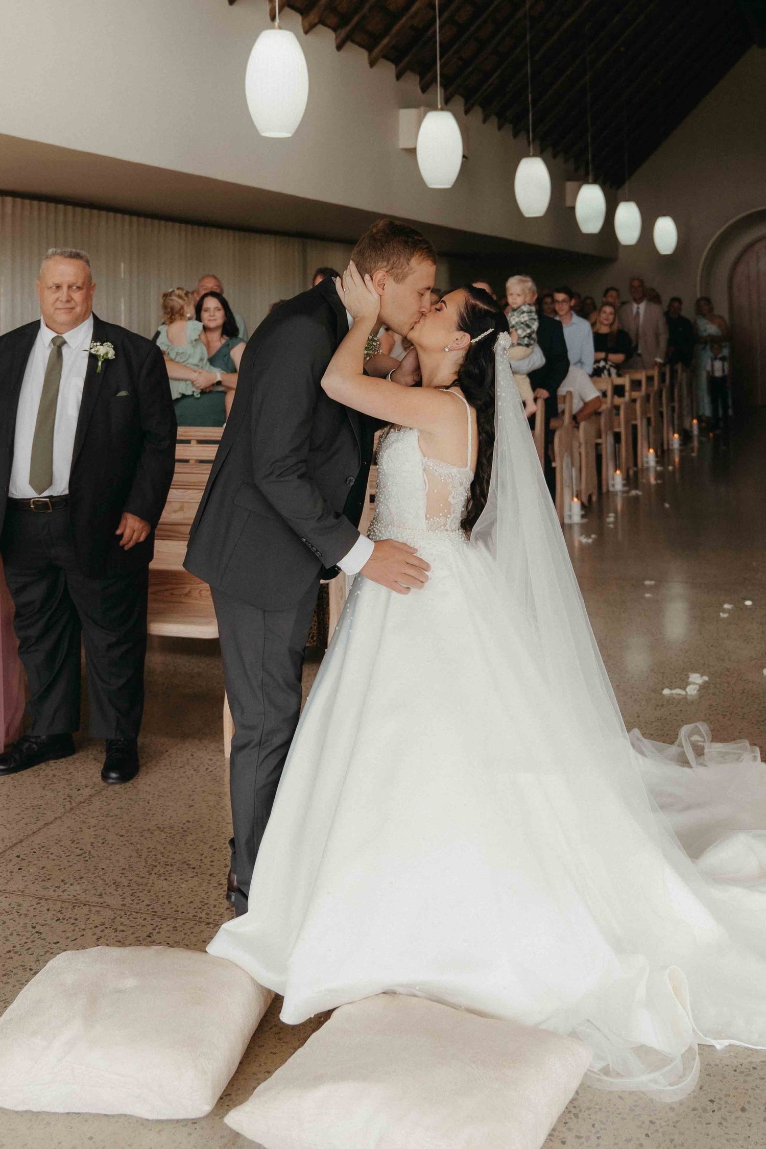 Bride and groom share first kiss at church altar, bride in flowing white gown with long train, guests seated in pews.