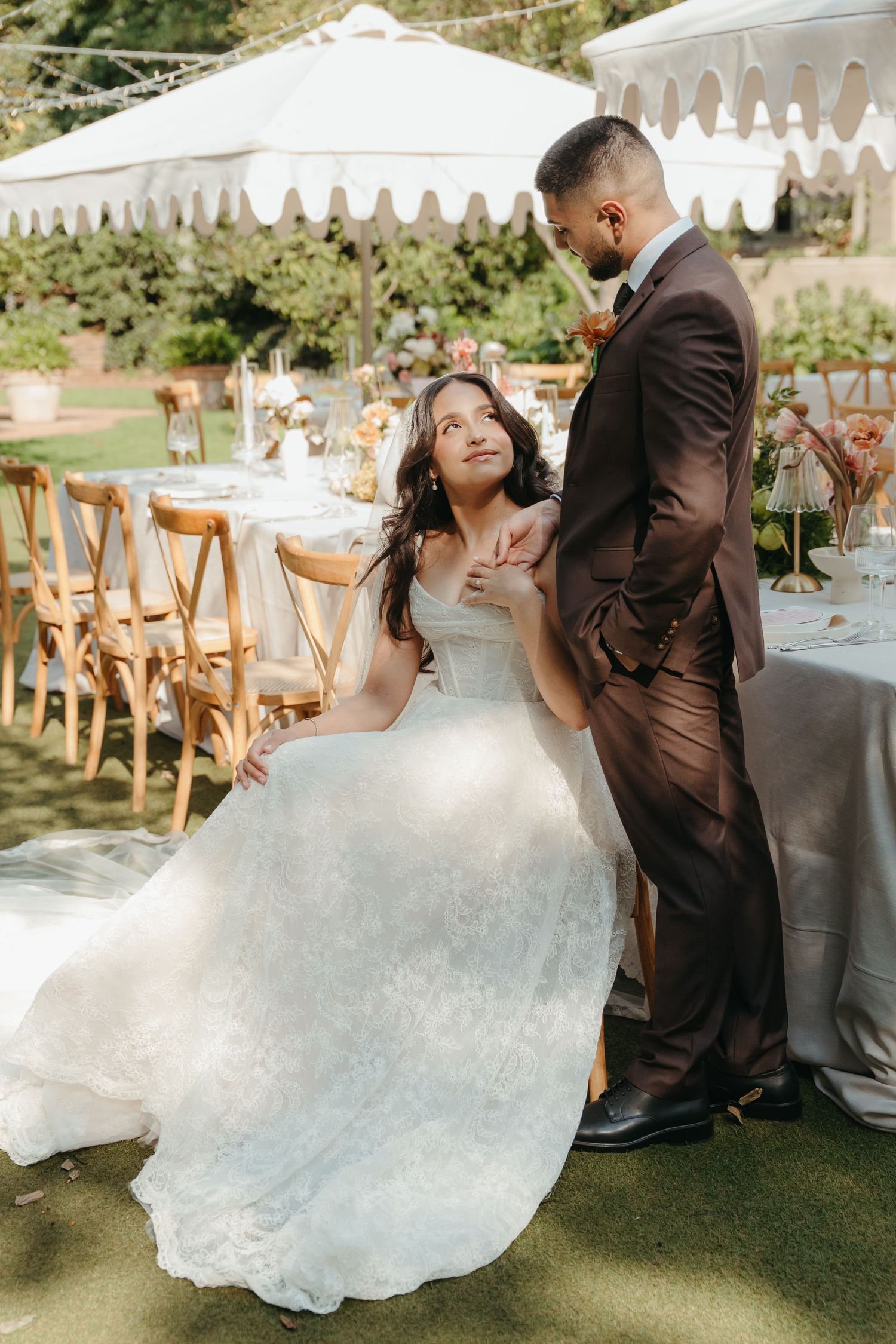 Bride in white lace gown sits gazing up at groom in brown suit holding hands at outdoor wedding reception.