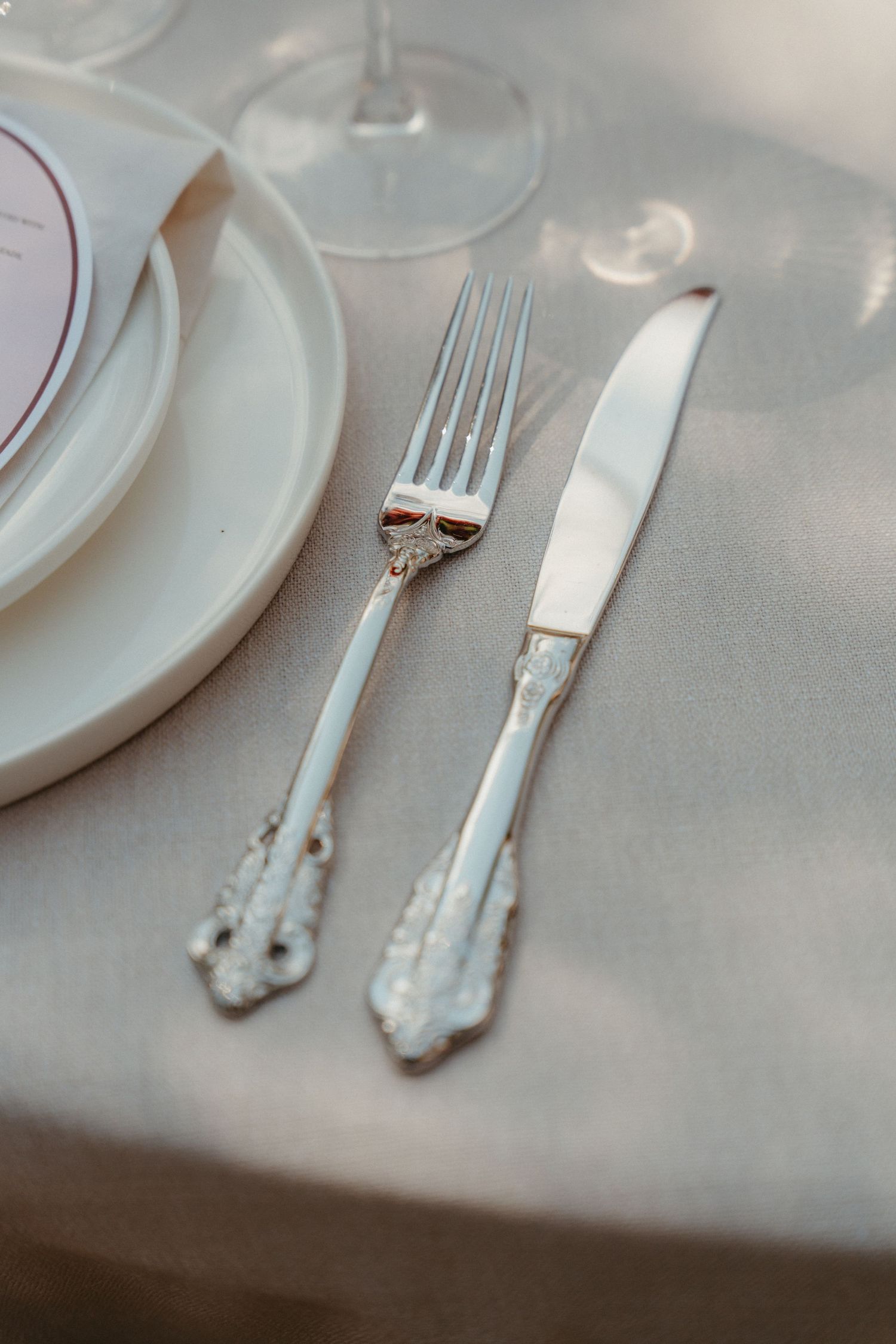 Elegant silver fork and knife with ornate handles on a linen tablecloth beside white plates at a formal place setting.