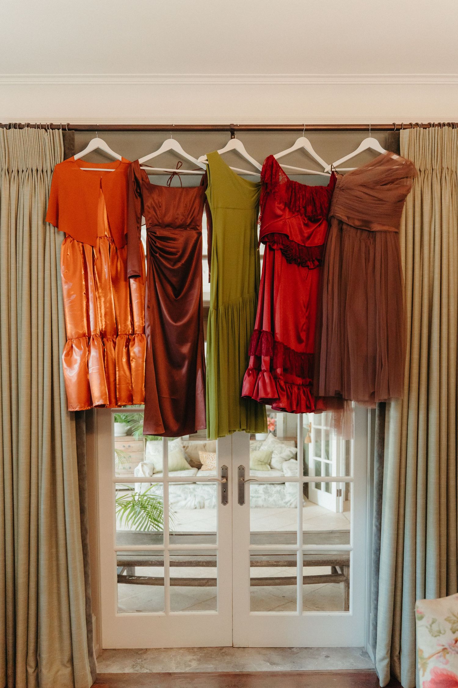 Five colorful bridesmaid dresses in orange, burgundy, olive, red, and mauve hanging on a curtain rod by French doors.