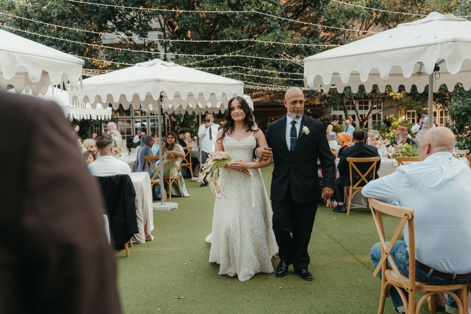 Bride walks down outdoor wedding aisle with escort, white umbrellas and string lights decorating the elegant garden venue.