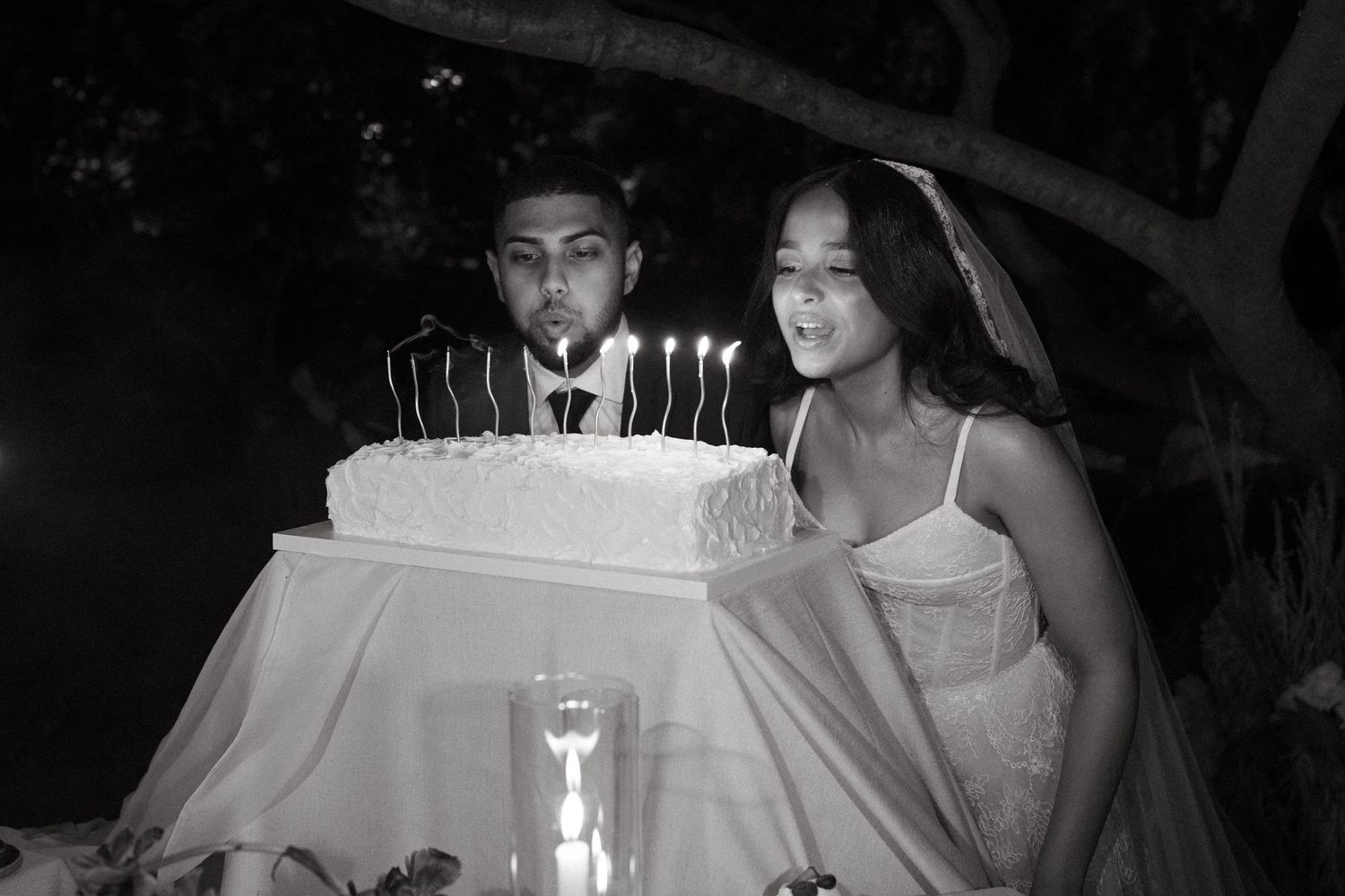 Black and white photo of a bride and groom blowing out candles on a wedding cake outdoors at night.
