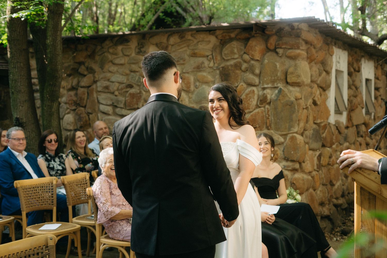 An outdoor wedding ceremony takes place in front of a rustic stone wall surrounded by trees and wooden seating.