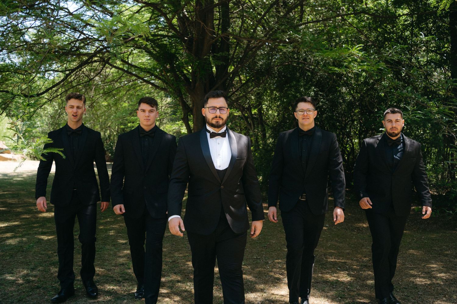 Five men in formal attire, with one in bow tie and tuxedo, walk together on a wooded garden path.