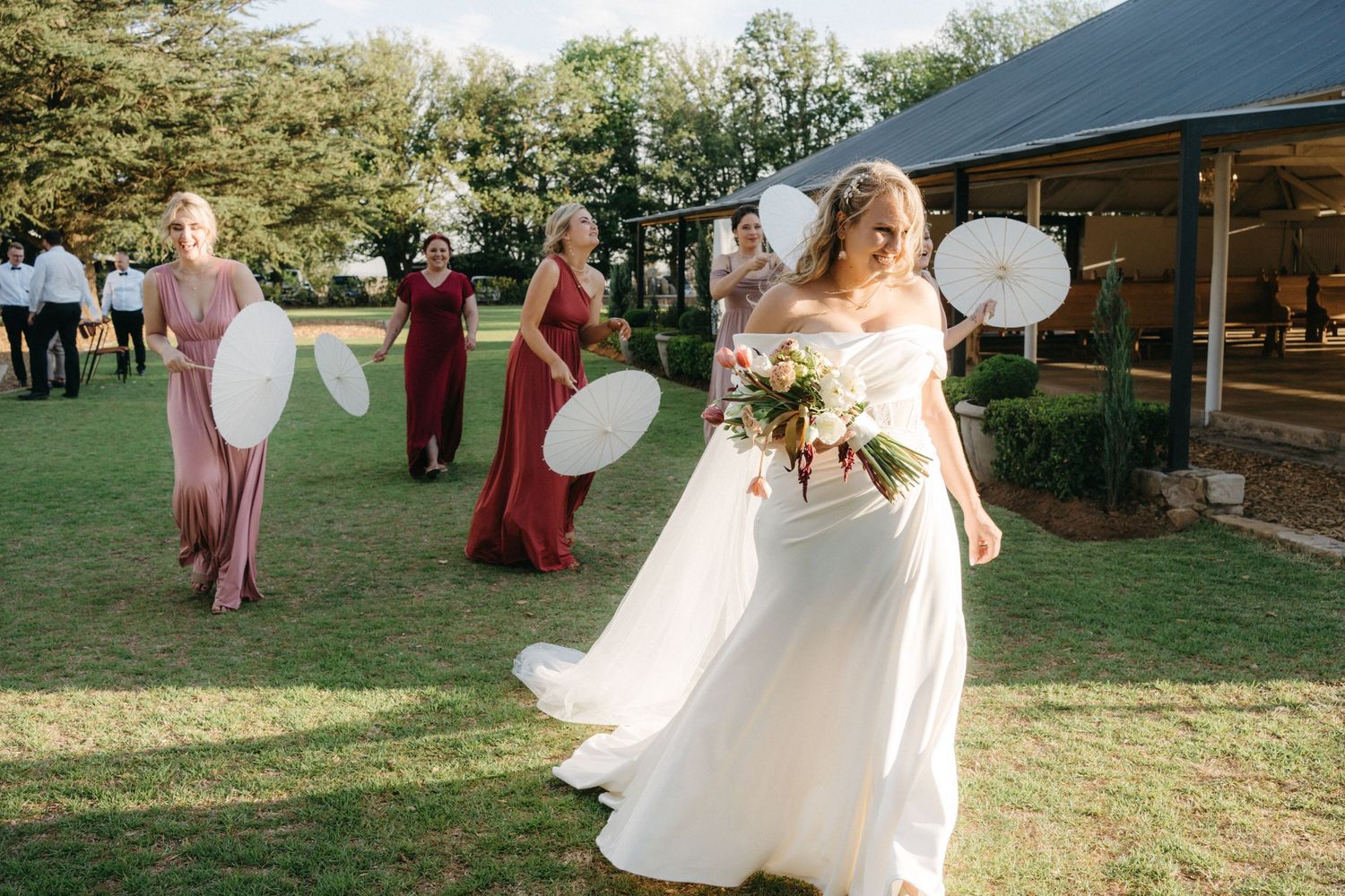 Wedding party walking across a lawn holding parasols at a rustic outdoor venue on a sunny day.