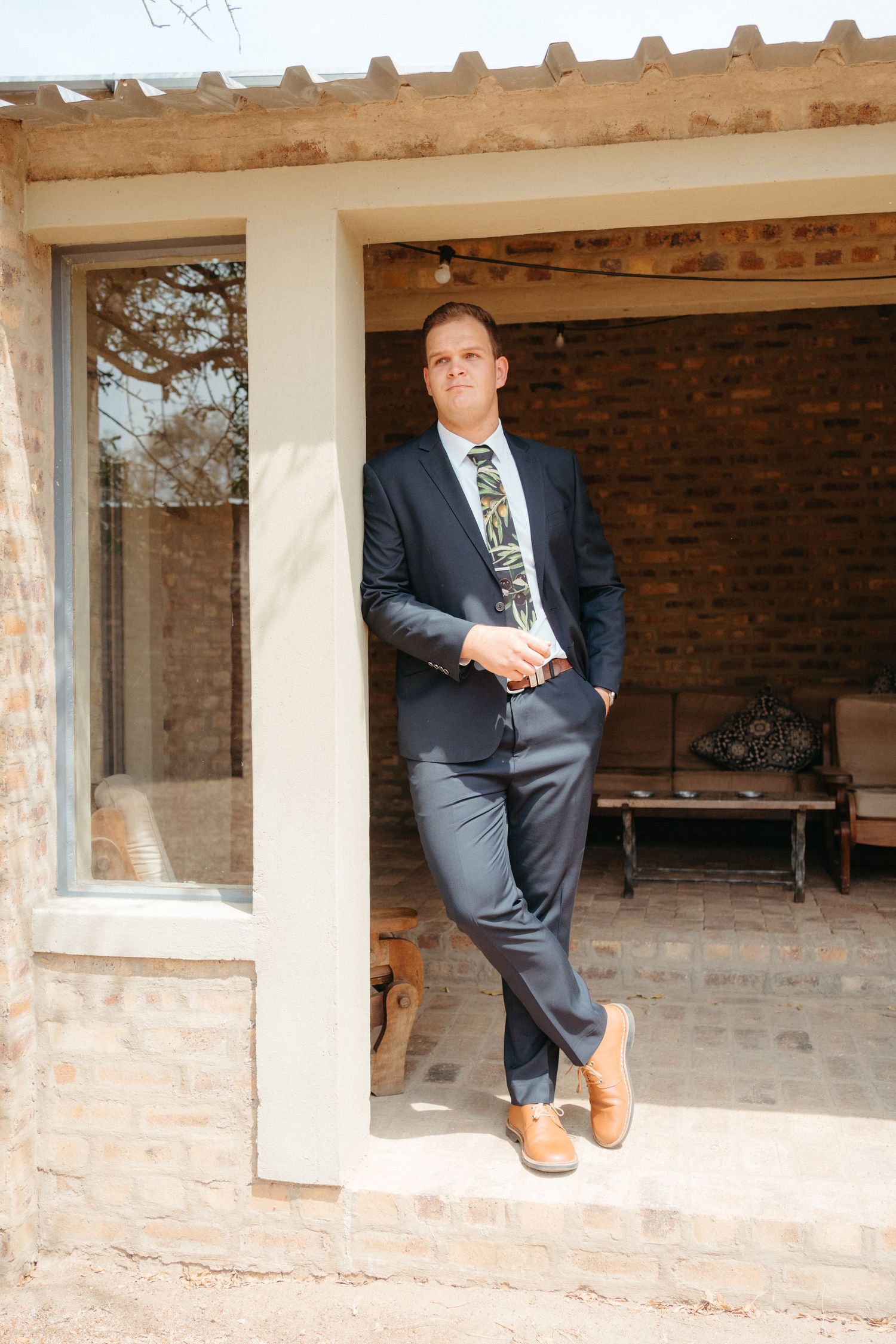 Formal portrait of person in navy suit leaning against rustic adobe building entrance.