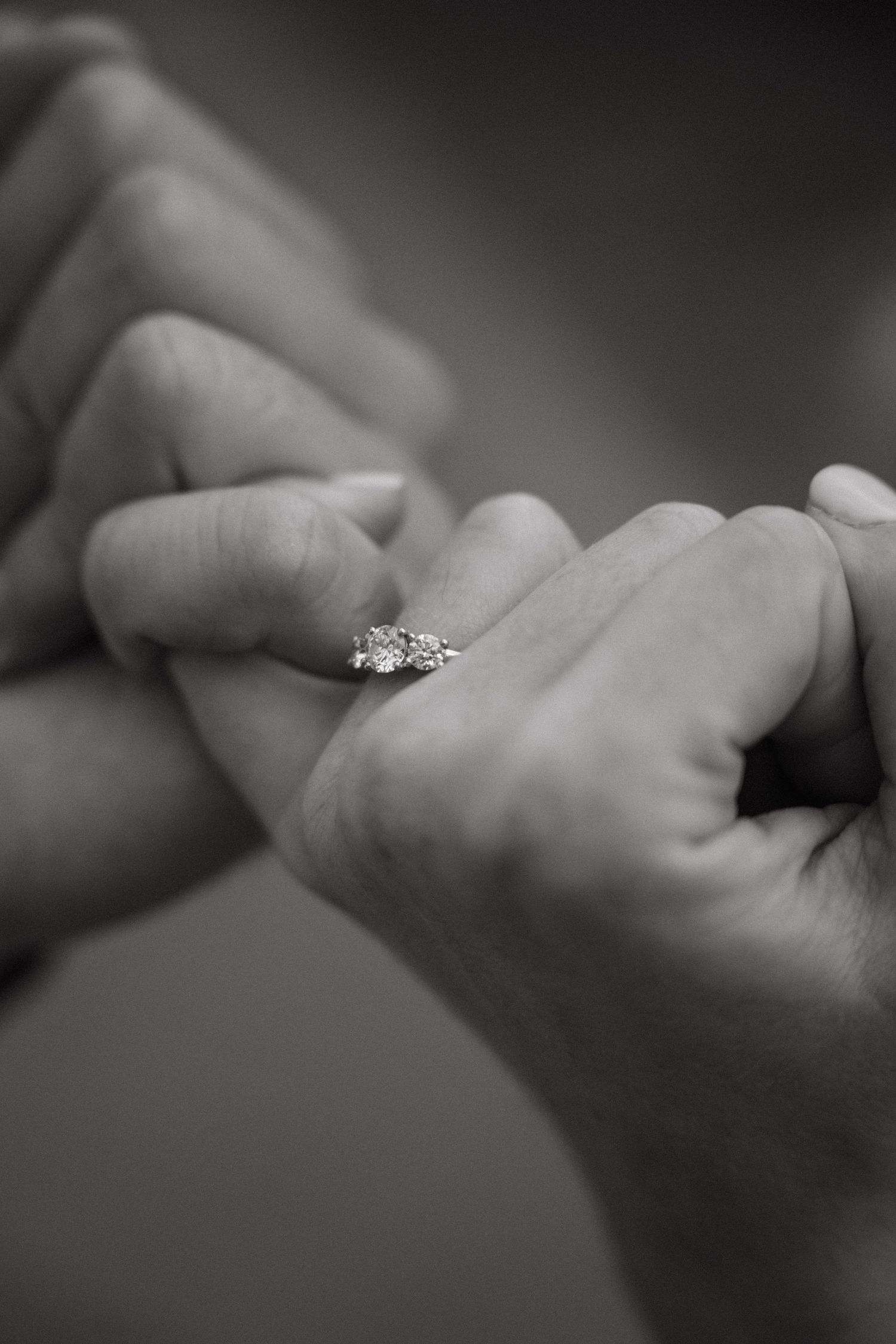 Black and white close-up of diamond engagement ring on woman's finger held by partner's hands.