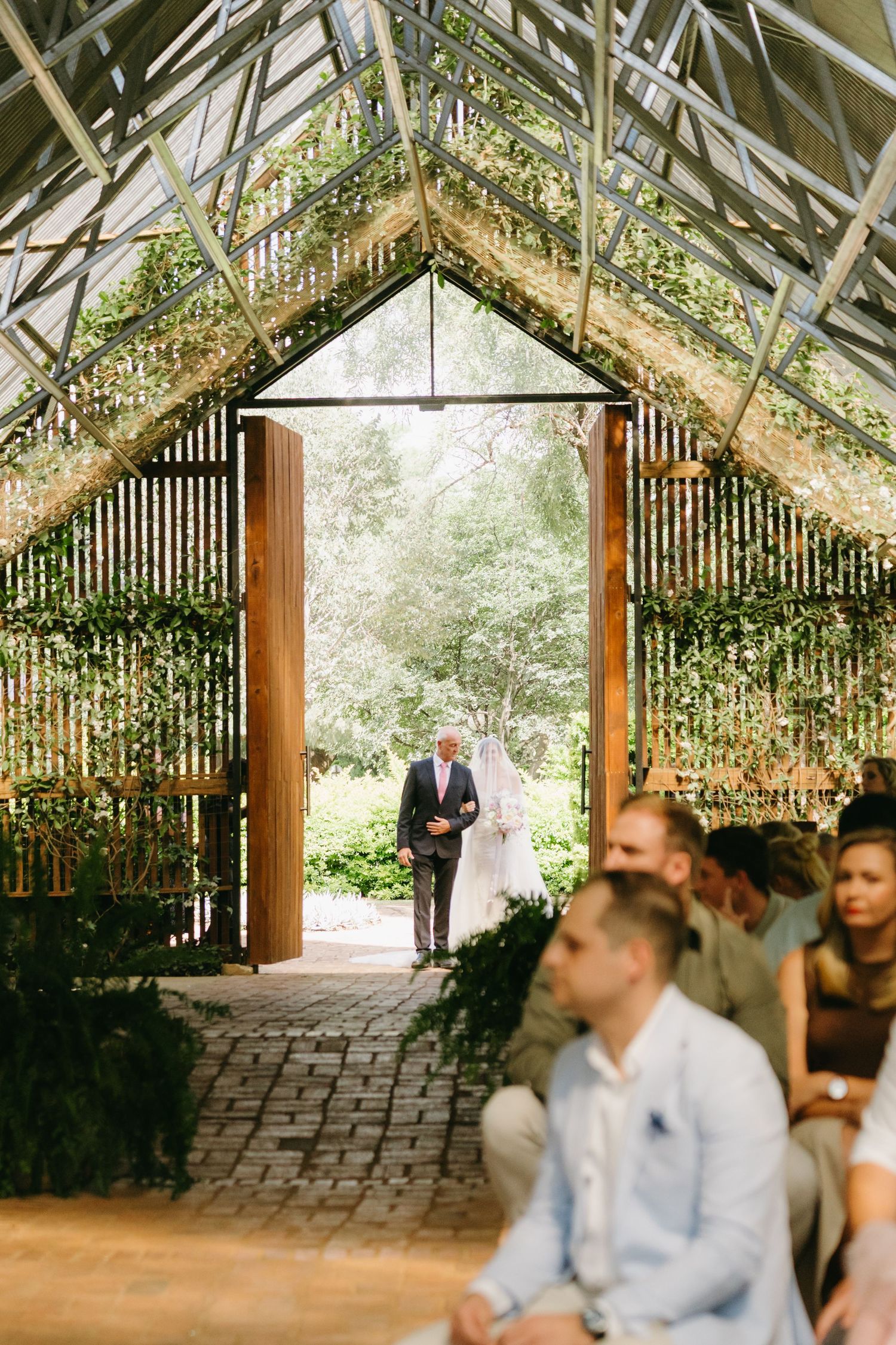 Wedding ceremony processional in greenhouse venue with natural light streaming through glass roof.