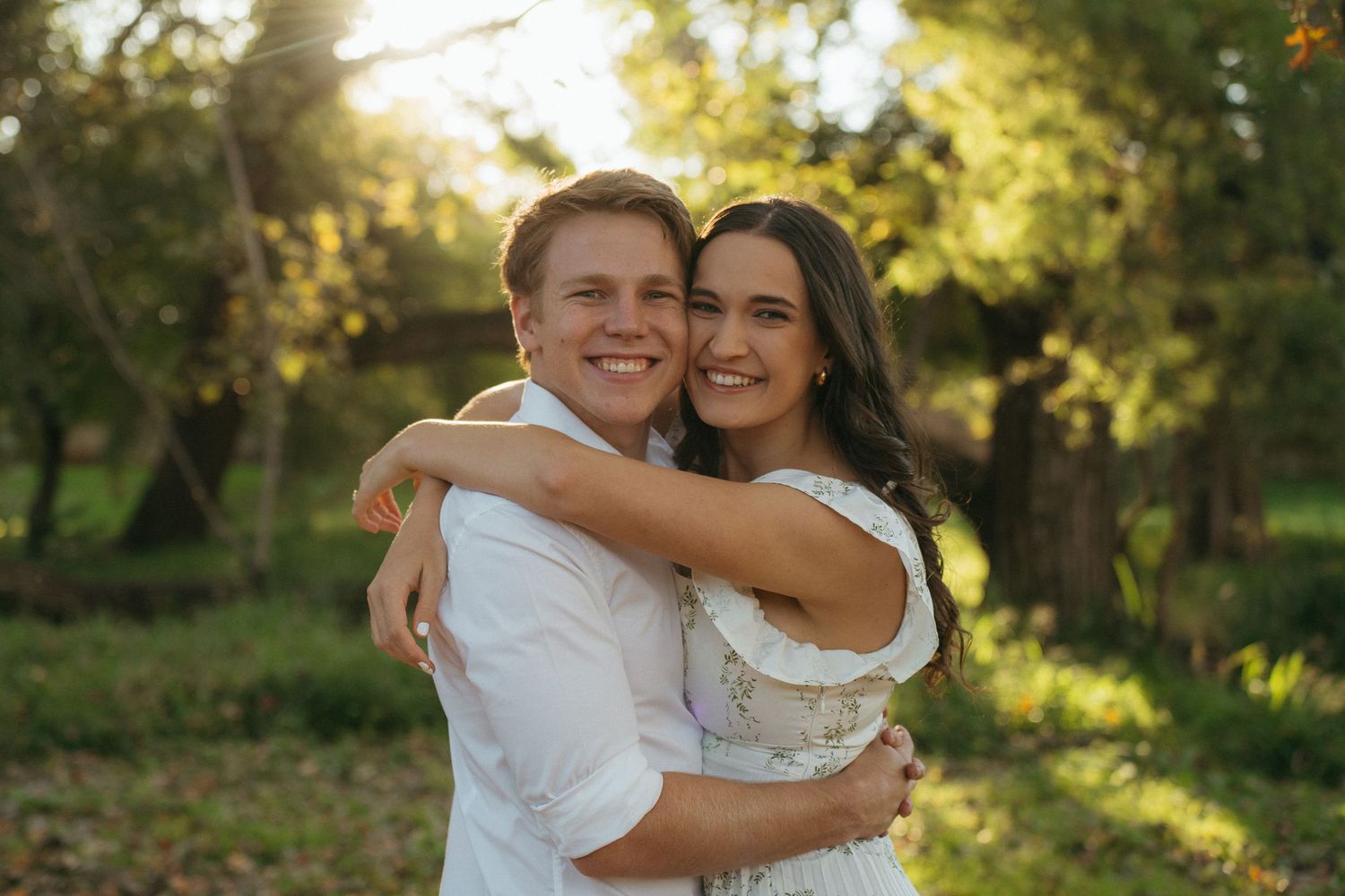 A happy couple in white outfits embrace while standing in a sunlit wooded area during golden hour.