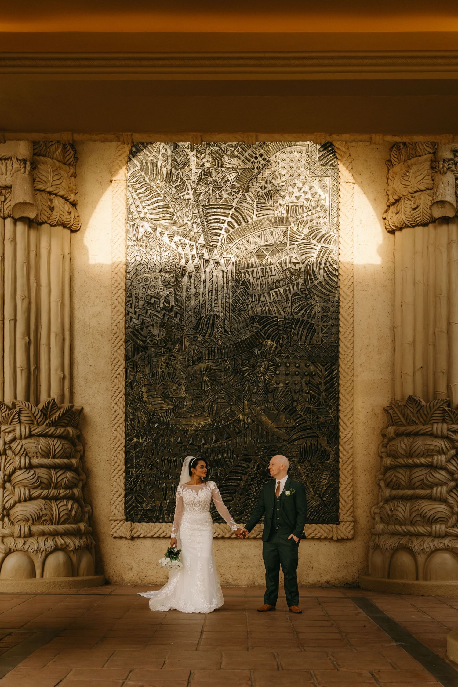 Wedding couple poses in front of illuminated mosaic wall between decorative stone pillars.