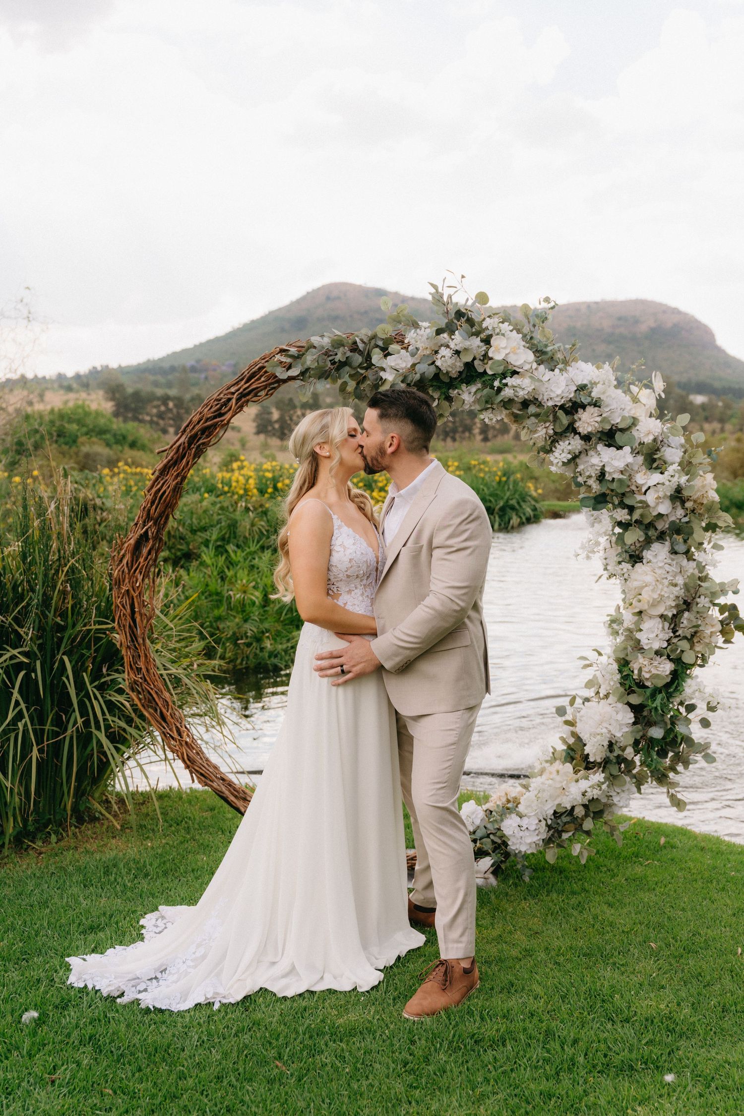 A wedding couple kisses under a floral arch with mountains and a lake in the background.