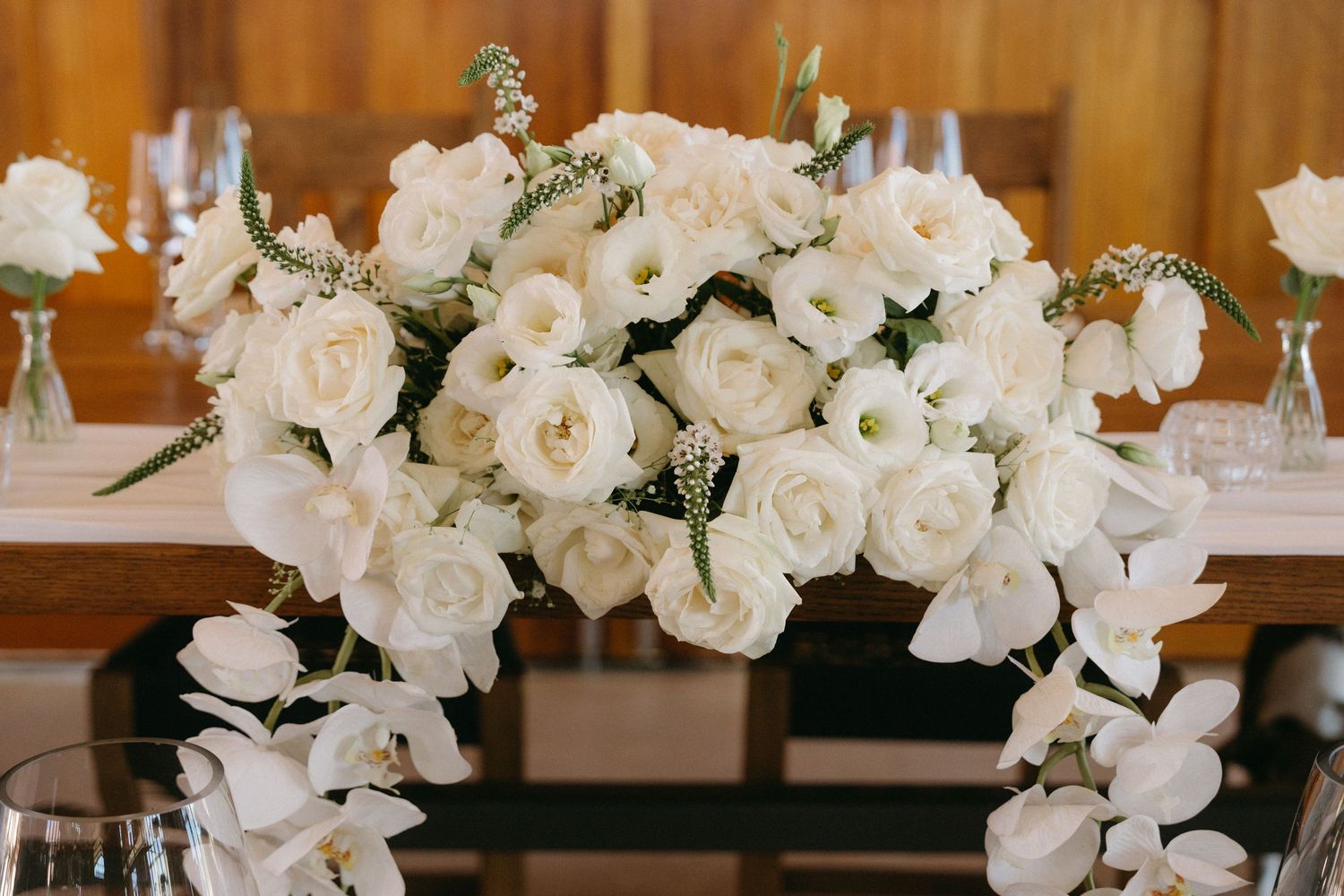 An elegant floral centerpiece of white roses and orchids arranged on a wooden table in rustic wedding decor.