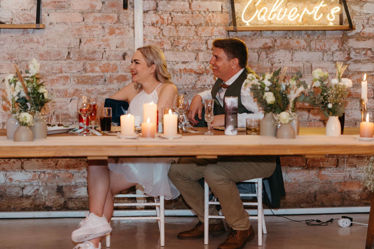 Wedding couple sits at a rustic reception table with brick walls and candlelight creating an intimate atmosphere.