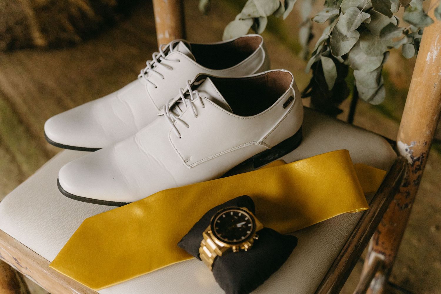 White dress shoes, gold watch, and yellow tie arranged on wooden surface for wedding day accessories.