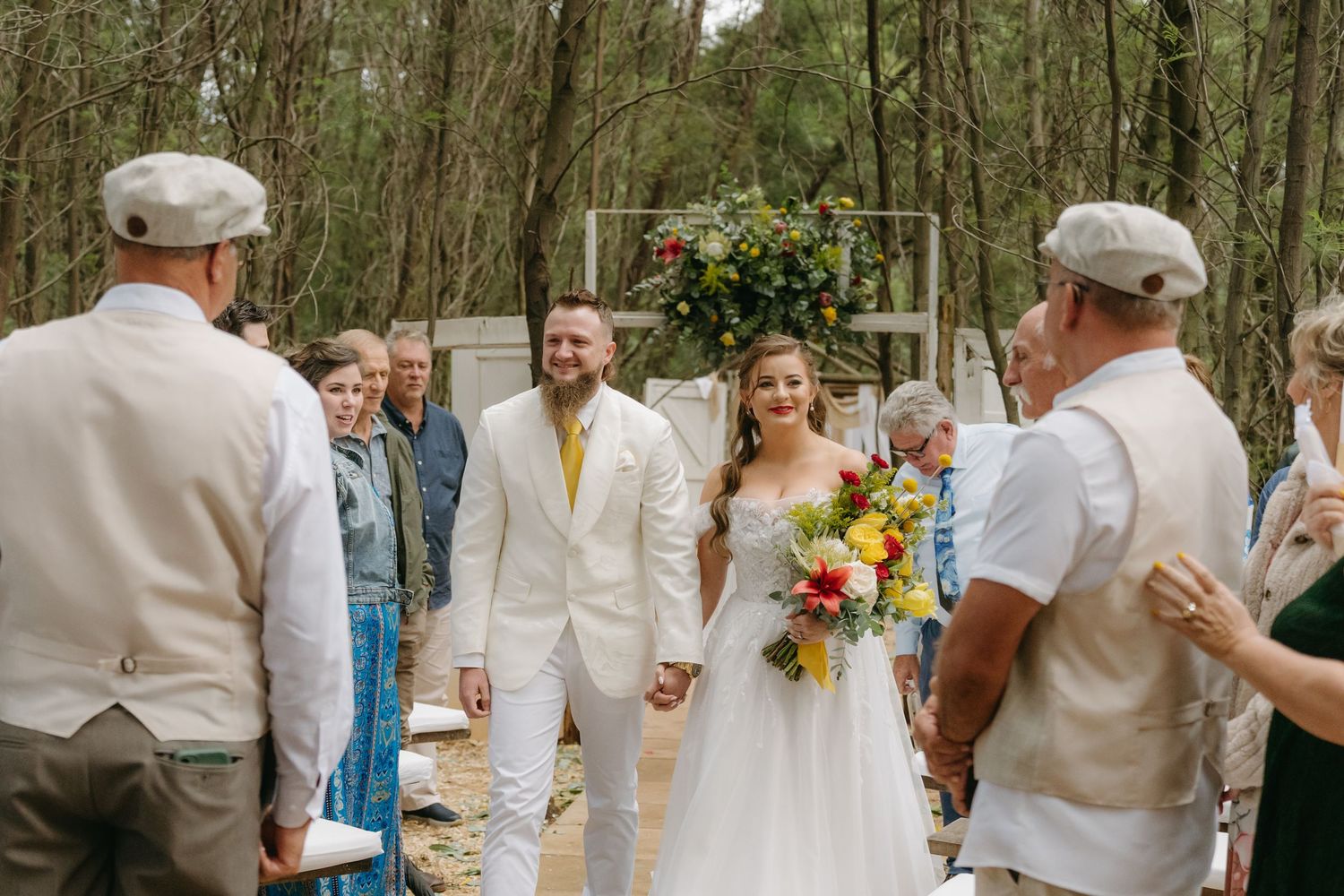 A couple walks down a forest path during their rustic outdoor wedding ceremony while guests look on.