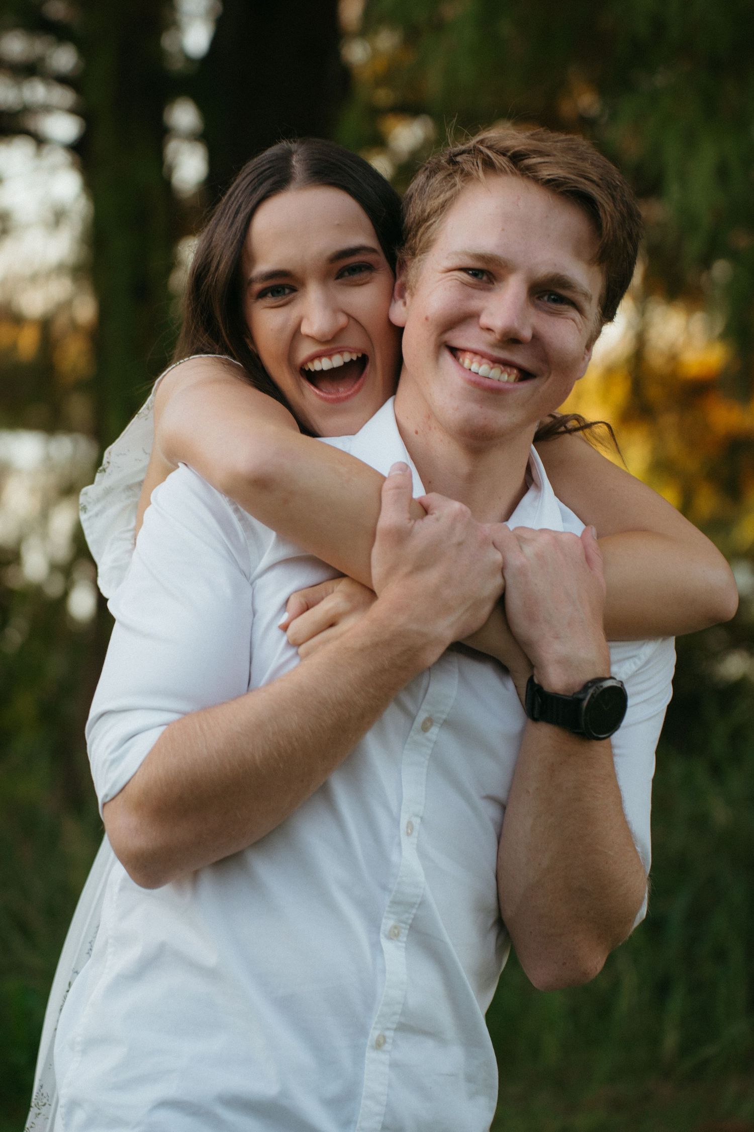Couple in white shirts embracing and laughing together outdoors with autumn foliage in the background.