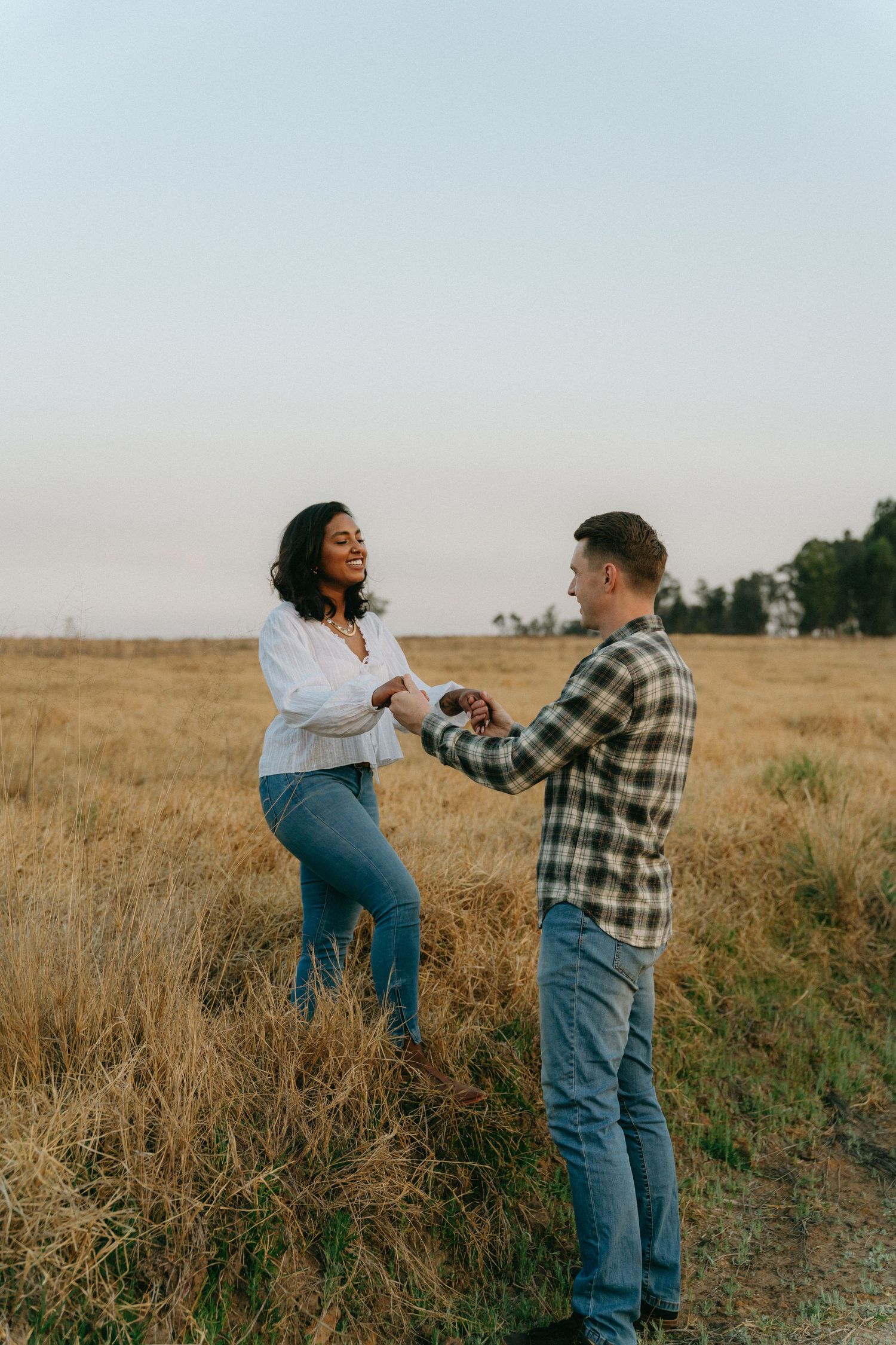 Playful couple dancing and twirling in a rustic field at dusk wearing casual attire.