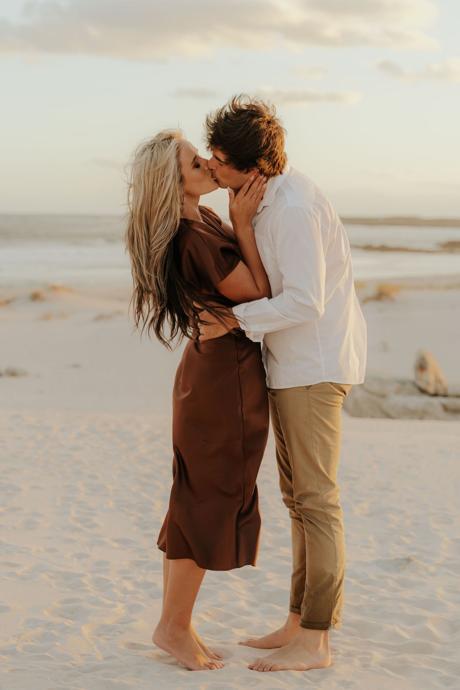 Couple sharing a romantic kiss on a sandy beach at sunset, woman in brown dress, man in white shirt.