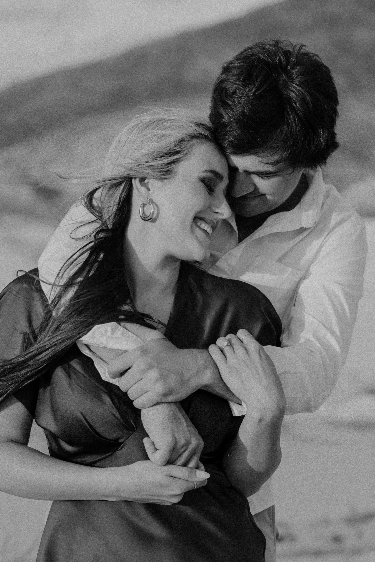 Black and white photo of a couple embracing romantically, woman smiling joyfully as man holds her close outdoors.