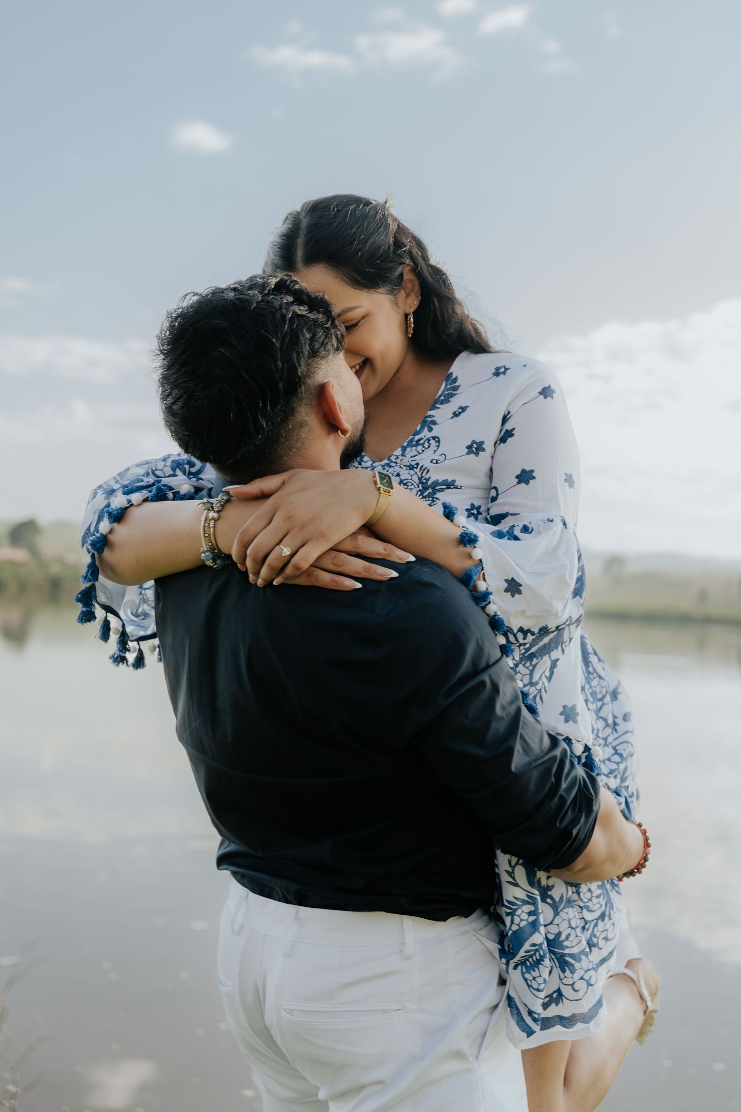 Romantic couple embracing by a lake, with her wearing a blue and white patterned dress and him in dark clothing.