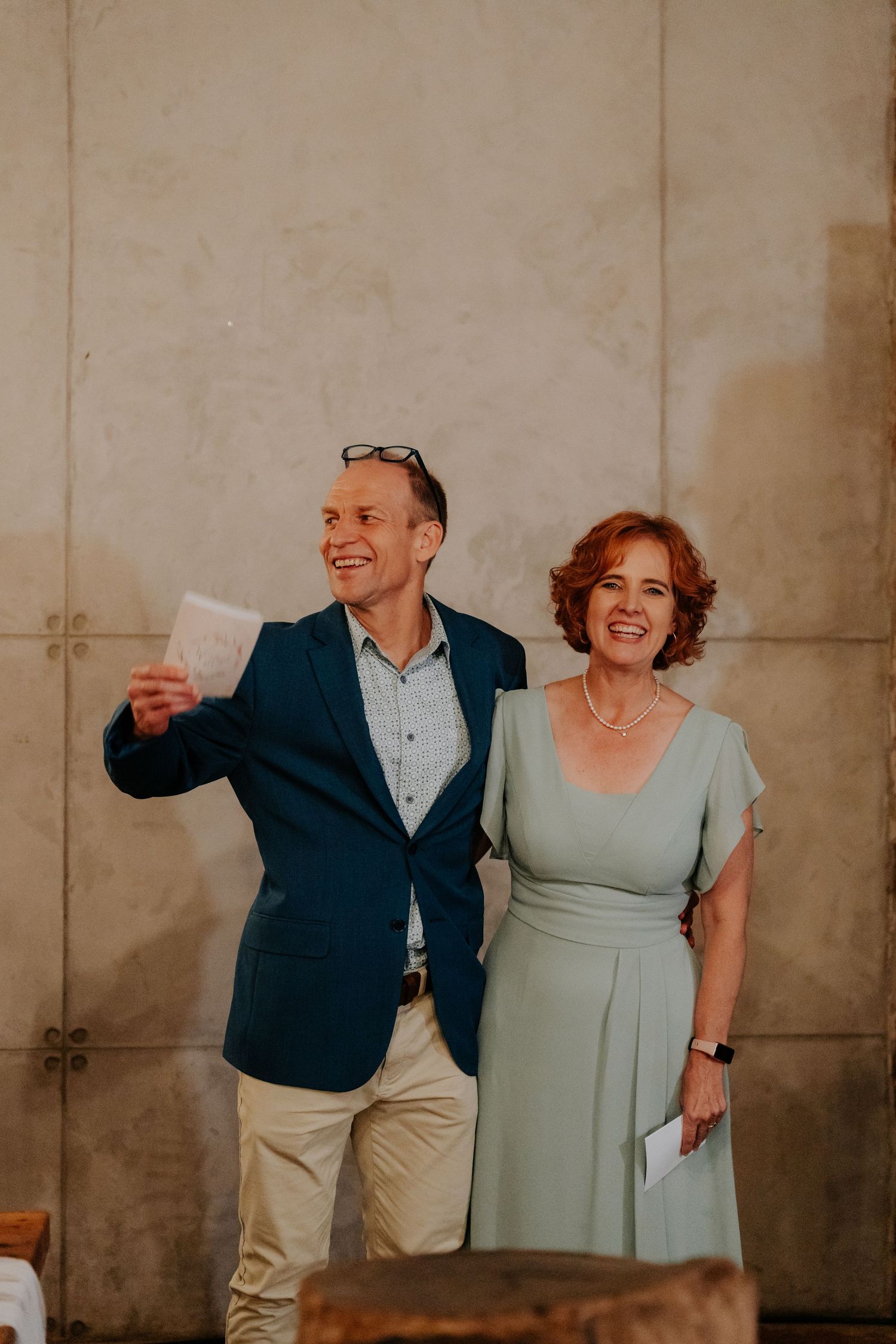 A group of people in casual attire laugh and share a joyful moment against a concrete wall background.
