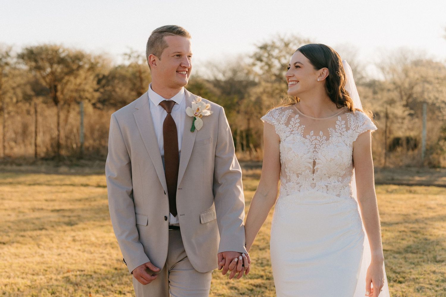 A bride and groom hold hands while standing in a golden sunlit field during their outdoor wedding ceremony.