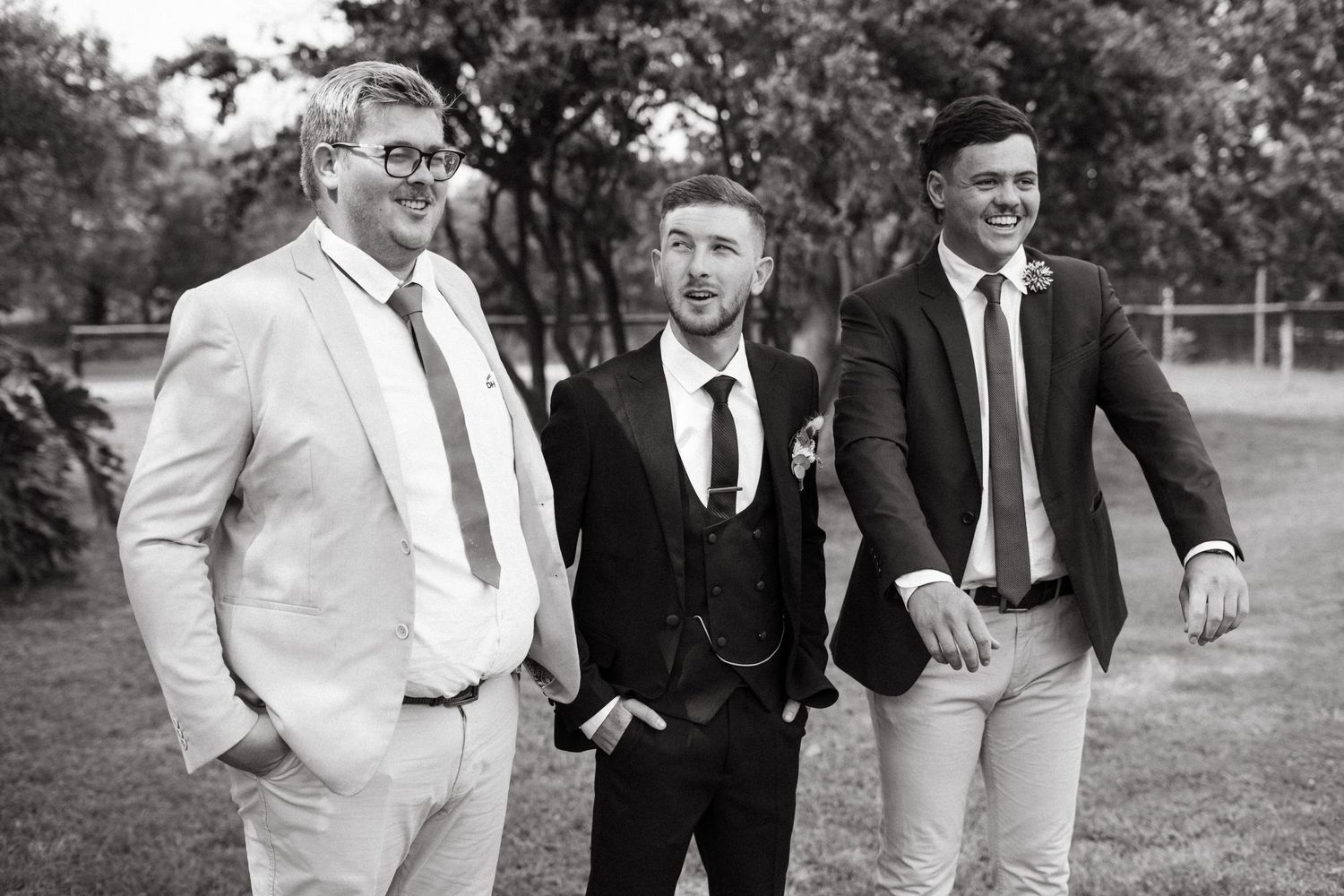 Three men in formalwear posing together outdoors for a black and white wedding photo.