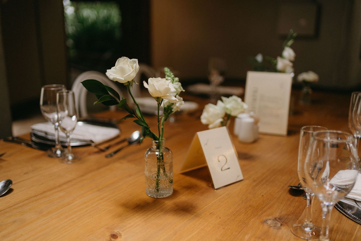 Elegant wedding reception table setting with white roses in glass vases and wine glasses on wooden table.