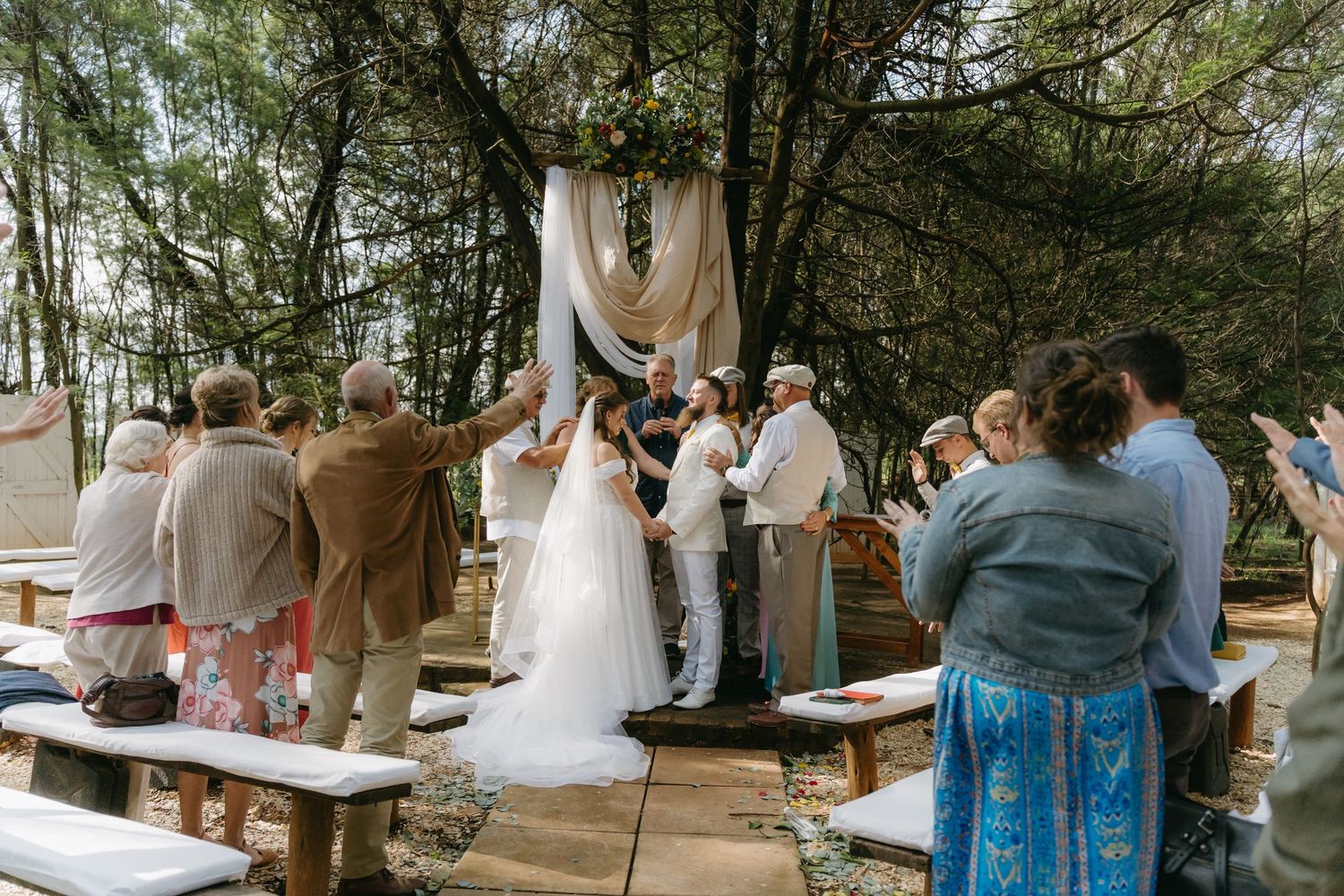 Outdoor forest wedding ceremony with guests seated on benches and draped arch backdrop.