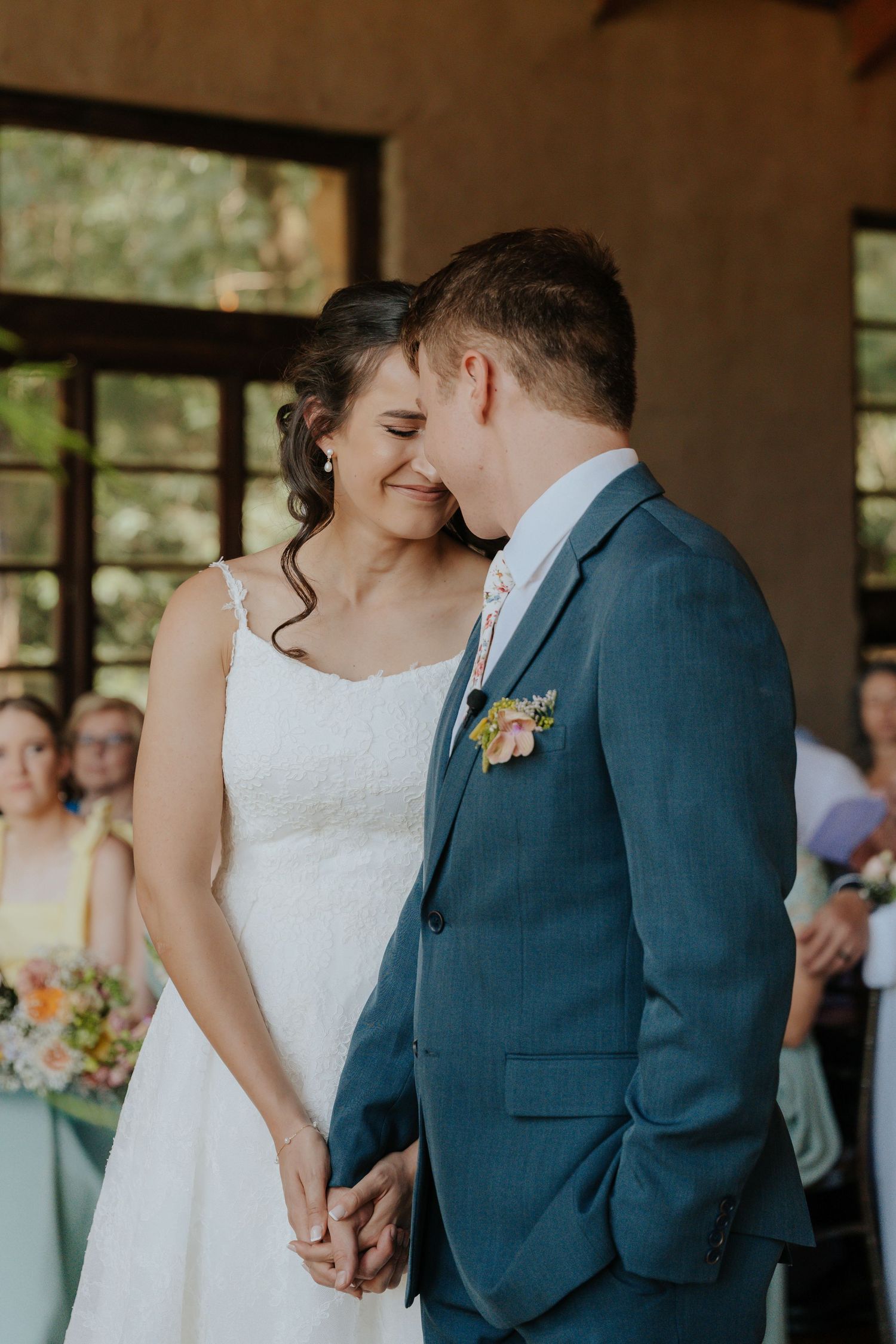 A tender moment between newlyweds in formal attire, with the bride in white lace and groom in navy blue suit.