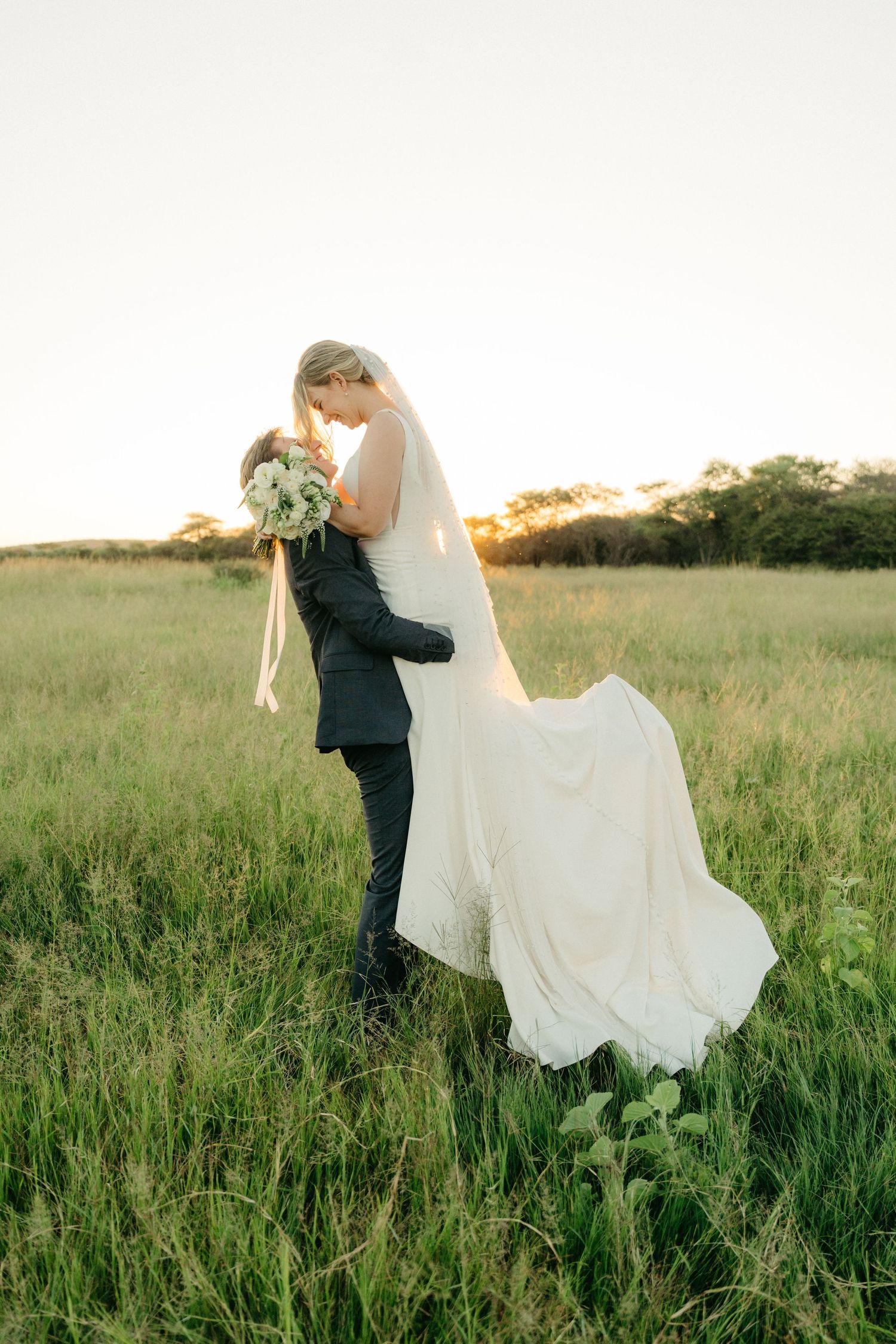 A newlywed couple embracing in a sunlit field with flowing white wedding dress and bouquet during golden hour.