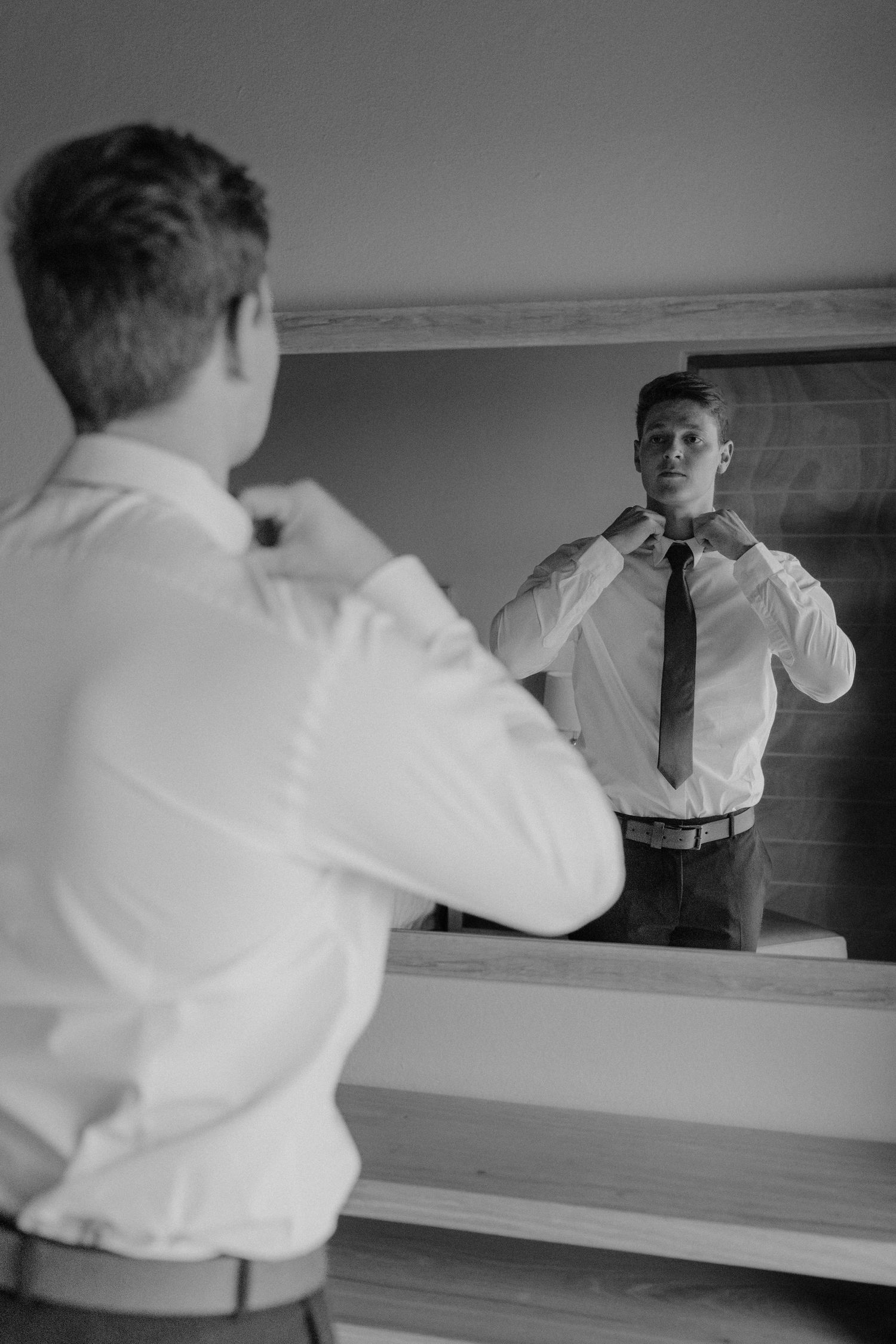 Black and white image of someone adjusting their tie while looking in a mirror before a formal event.