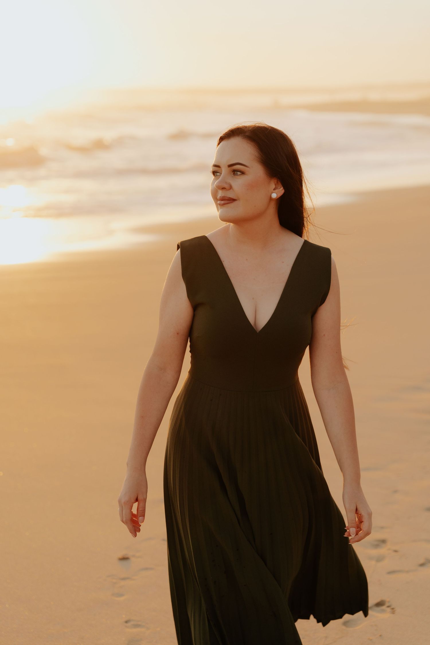 A person in a black dress walks along the beach at sunset creating a silhouette against the golden sky.