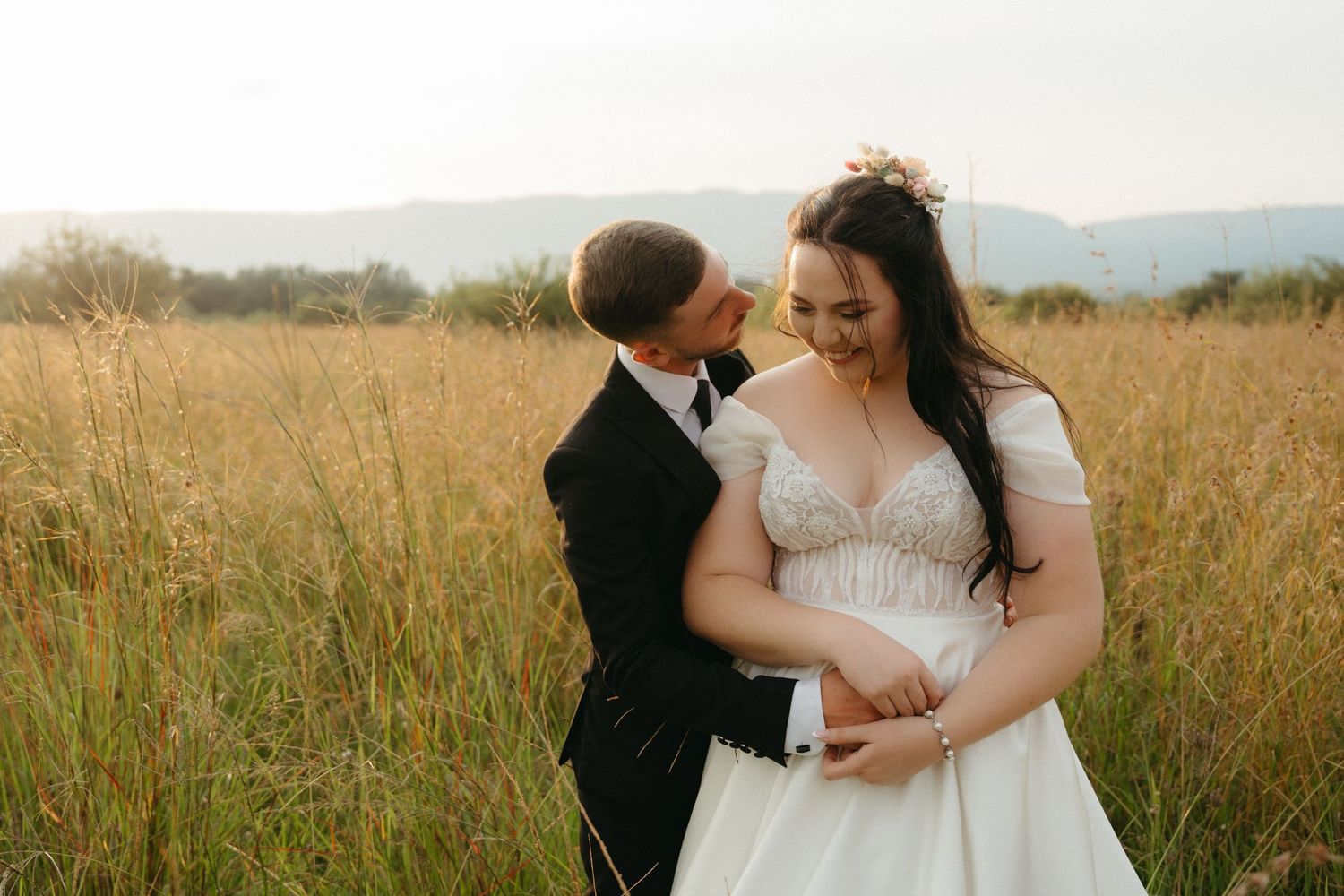 A romantic couple shares an intimate moment in a golden field at sunset, surrounded by tall grass and mountains.