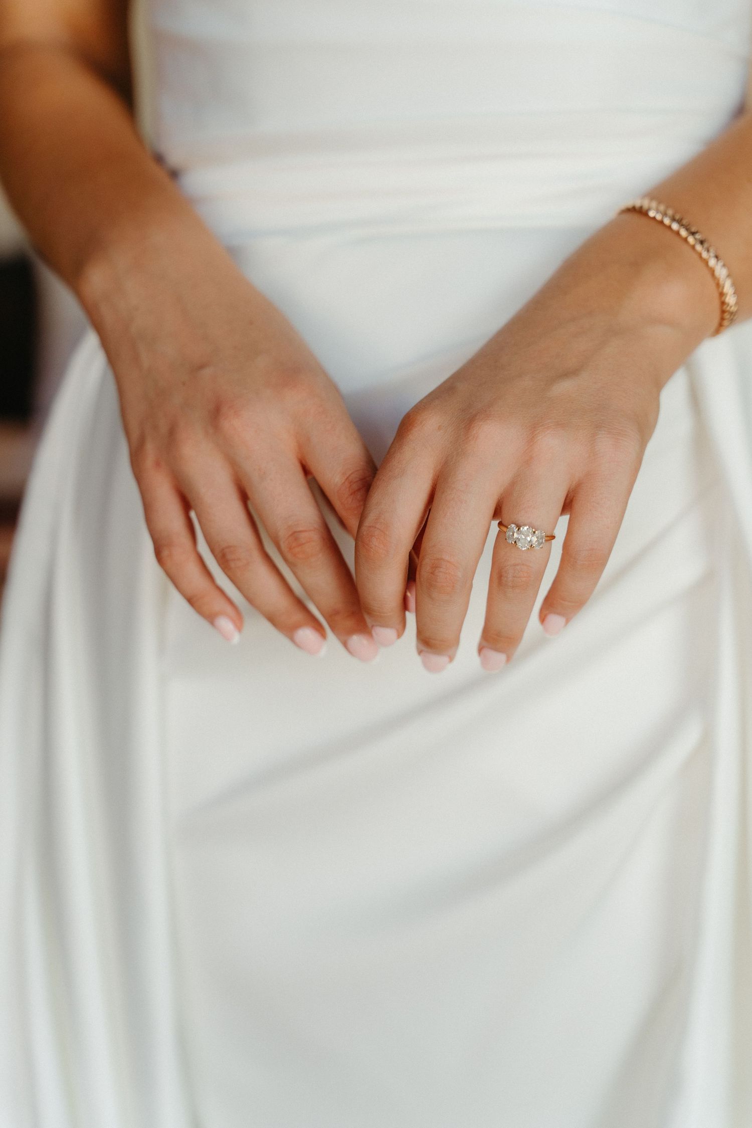 Close-up view of hands resting on a white wedding dress showing delicate jewelry and engagement ring details.