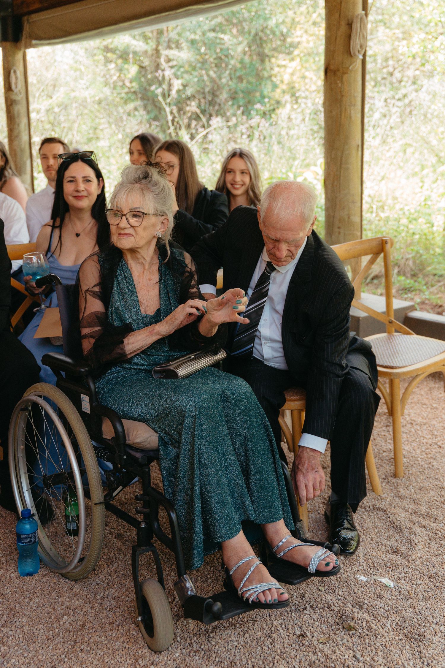 A group sits together in an outdoor covered area, with someone in a wheelchair and others in formal attire.
