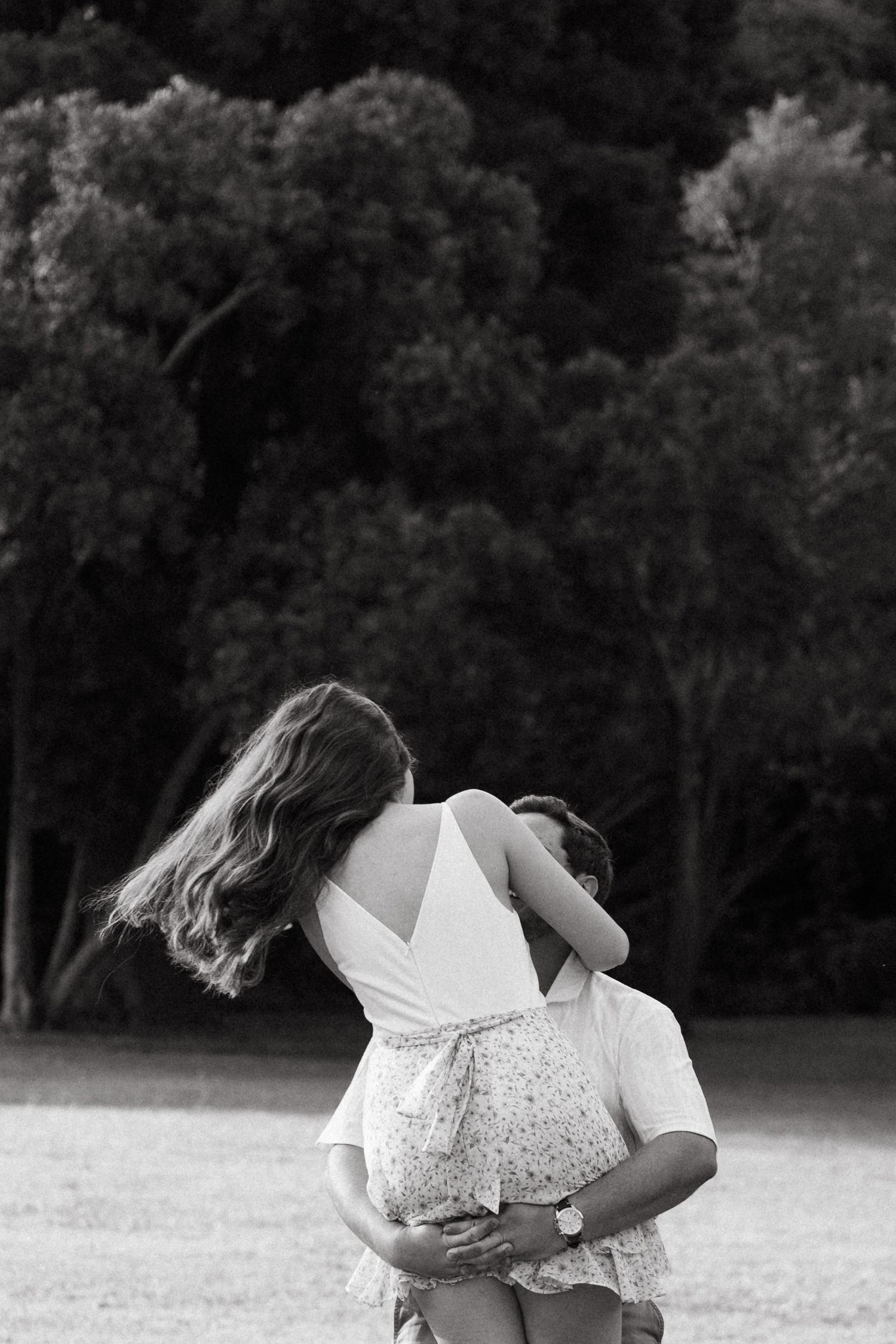 Black and white photo of a couple embracing in a park, woman lifted by man with trees in background.