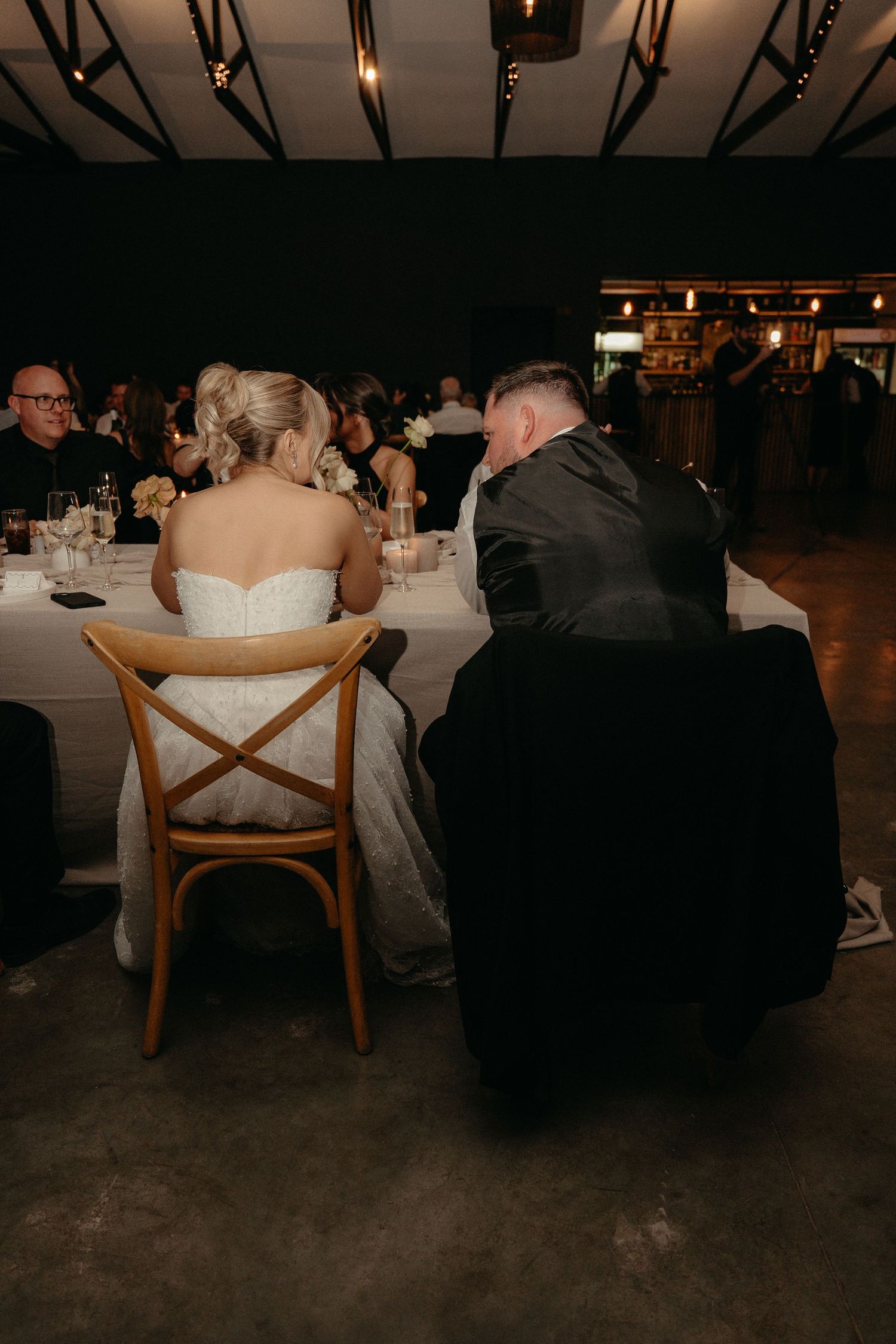 Wedding guests seated at elegant dining tables in a dimly lit reception venue with wooden cross-back chairs.