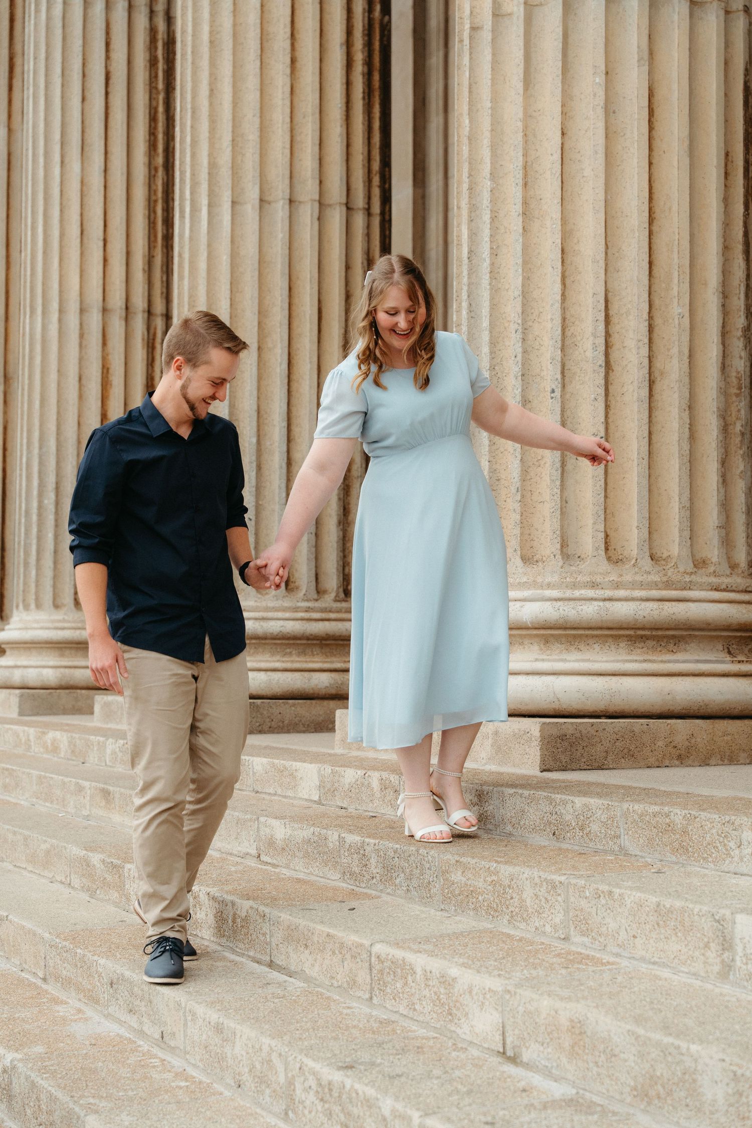 A couple holding hands while walking down stone steps in front of classical columns, with her wearing a light blue dress.