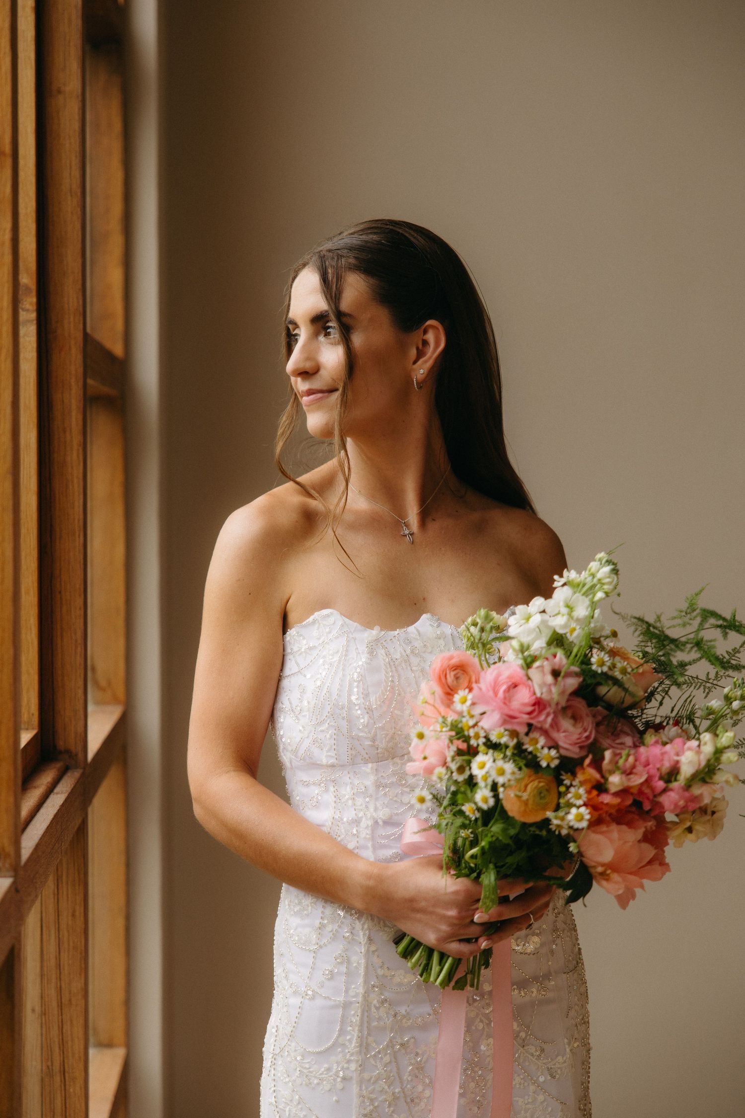 Bride in strapless lace gown holding colorful pink floral bouquet with ribbon, standing by window.