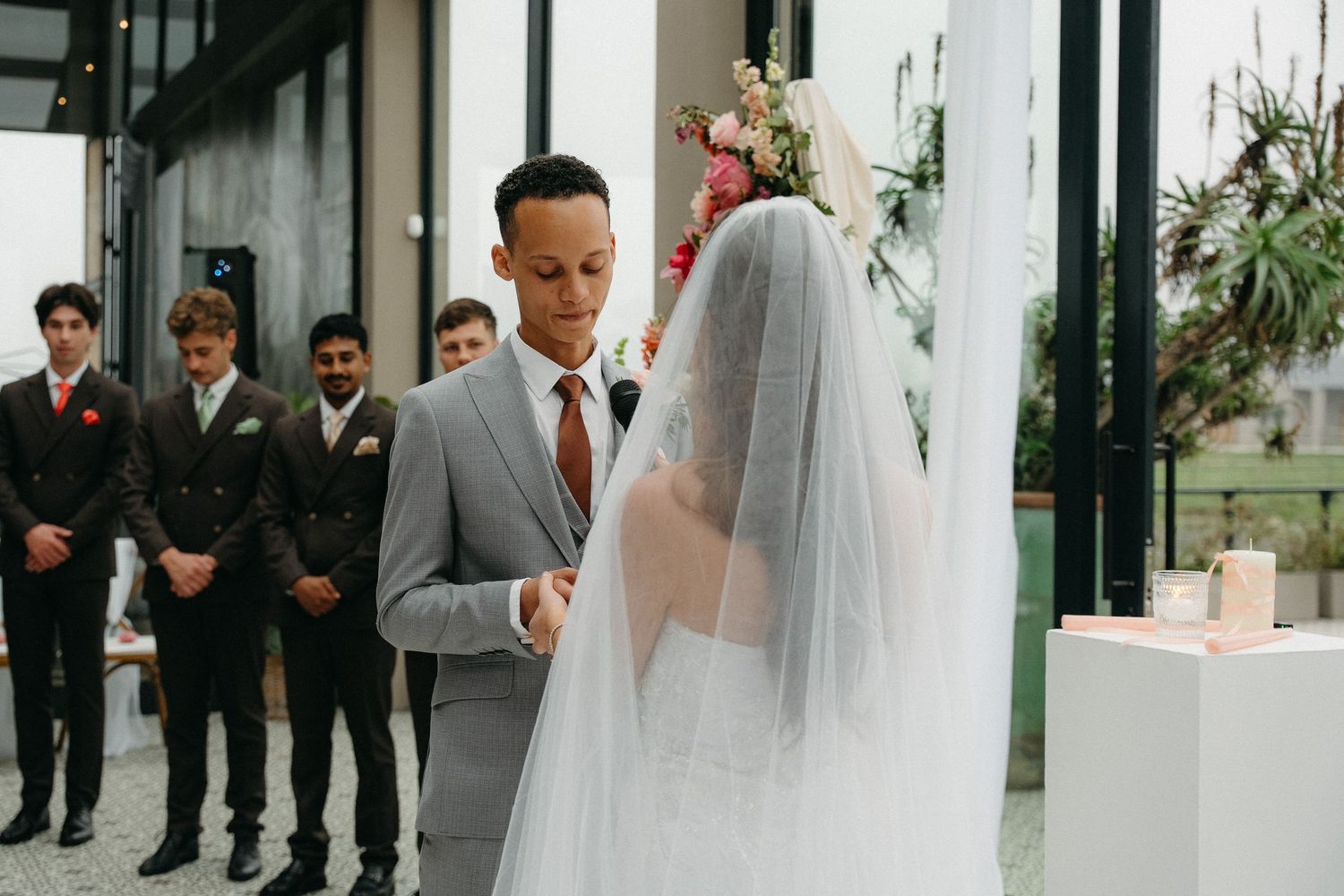 Groom in gray suit exchanges vows with veiled bride at elegant indoor wedding ceremony with groomsmen standing behind.