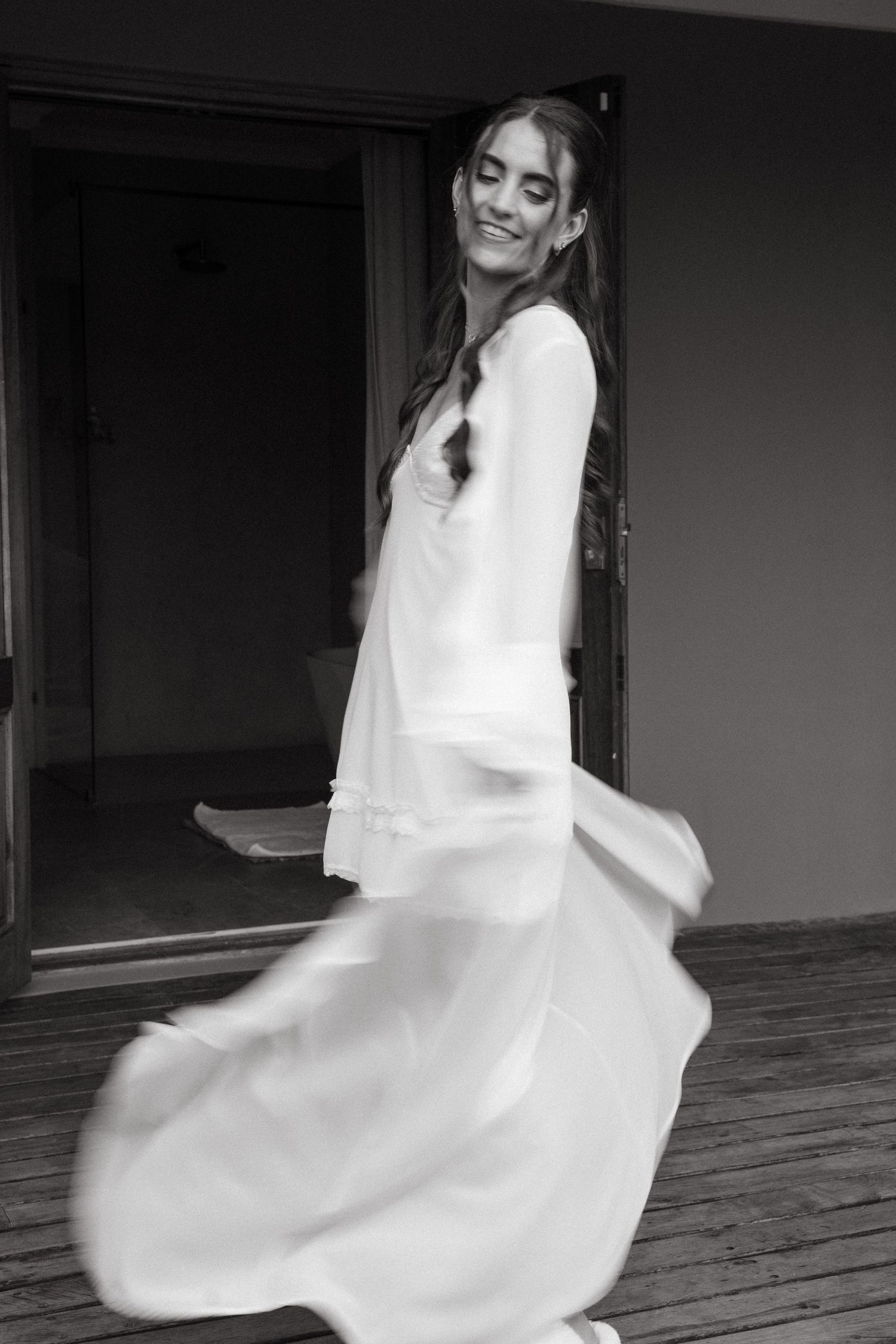 Black and white photo of a smiling bride in a flowing white robe twirling on a wooden deck.