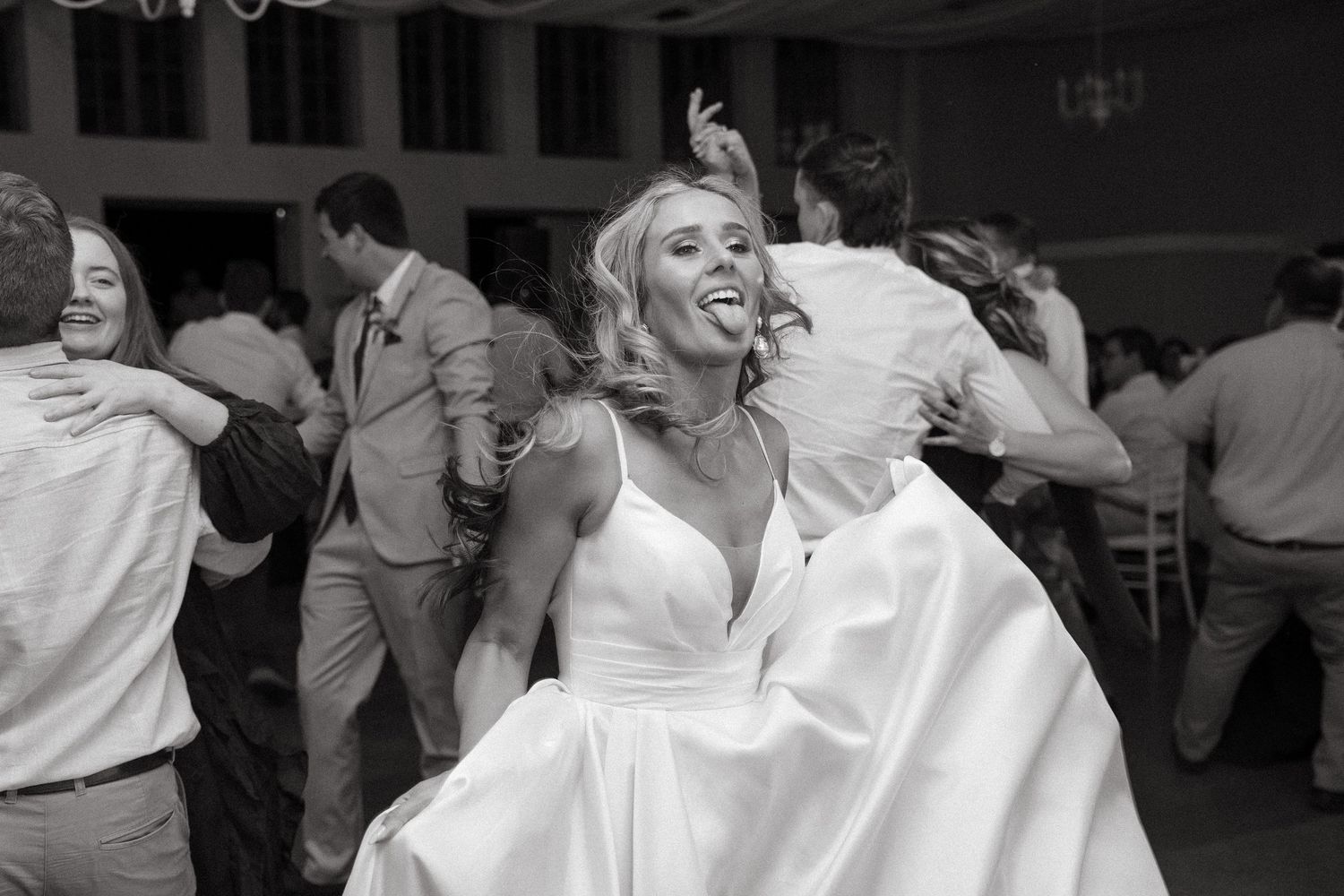 Joyful bride in a white wedding gown sticking out her tongue while dancing with guests on the dance floor at a wedding reception.